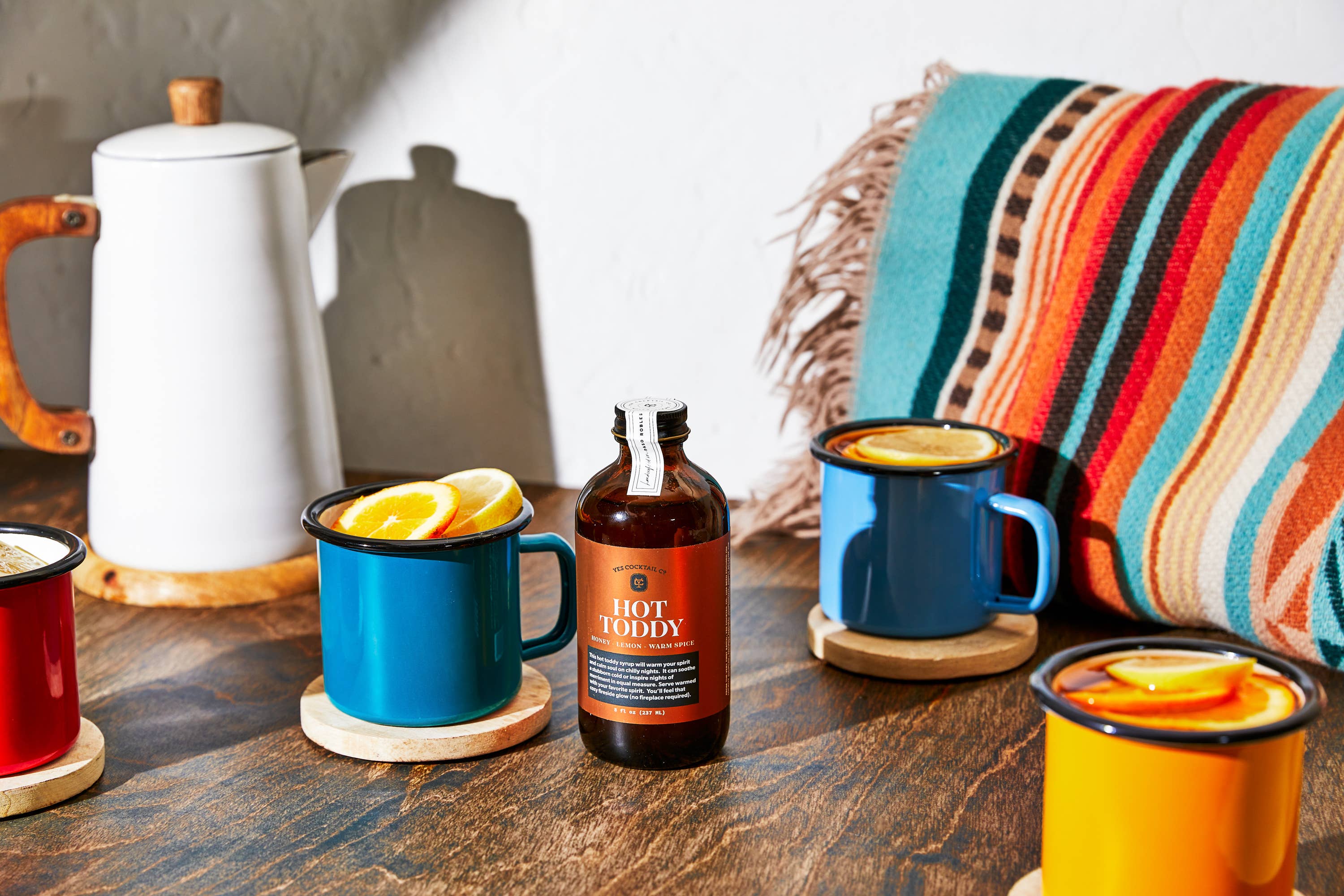 A cozy setup with a white teapot, a bottle labeled 'Hot Toddy,' and four colorful mugs (red, blue, yellow, and another blue) filled with a beverage garnished with orange slices. The mugs are placed on wooden coasters on a wooden table. A colorful striped blanket in the background adds to the warm and inviting atmosphere, suggesting a relaxing and comforting setting for enjoying a hot toddy on a chilly day.