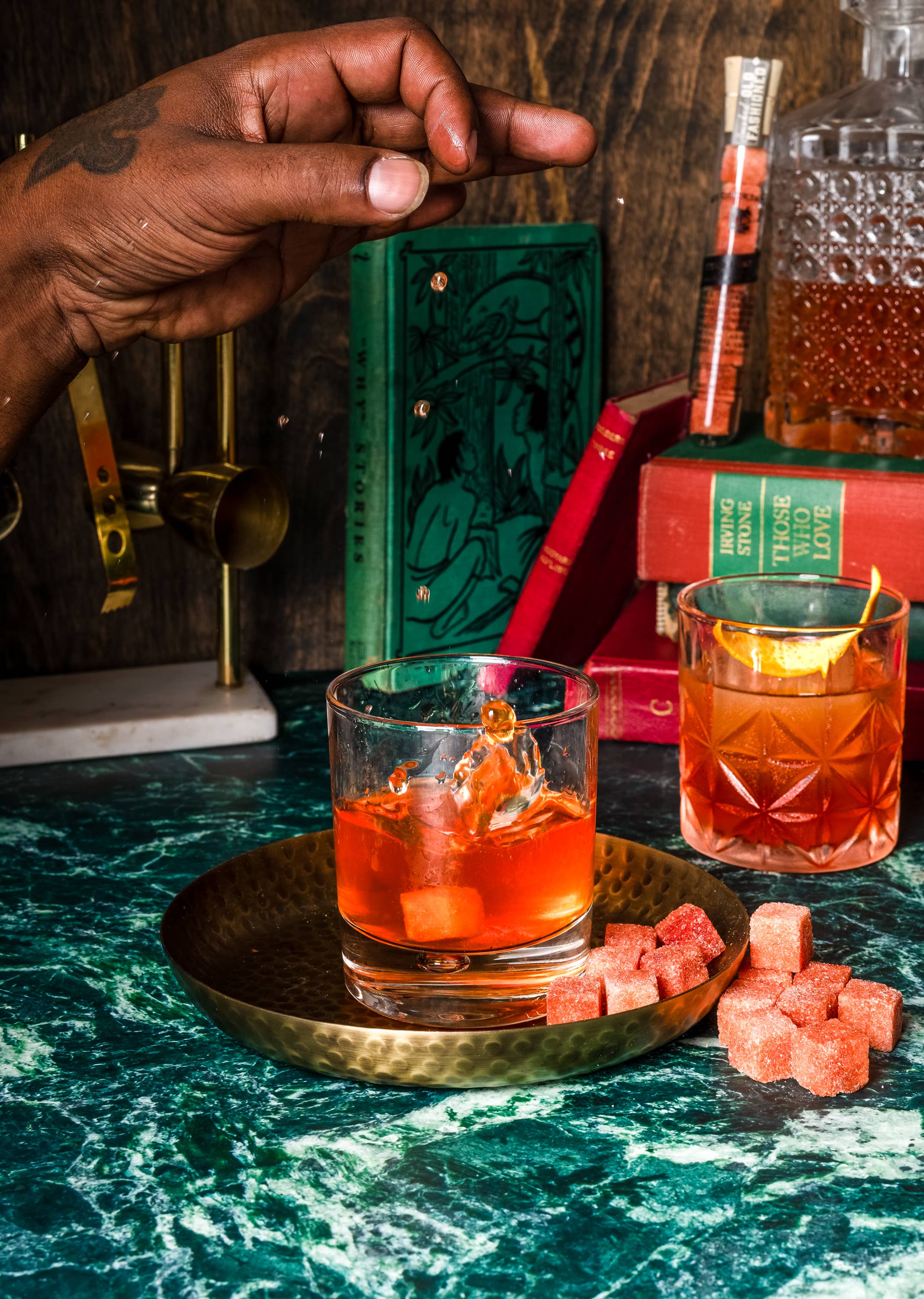 A hand sprinkles a garnish over a glass of orange-colored cocktail placed on a brass plate. Next to the glass are red sugar cubes, likely Ace Rivington Old Fashioned Bitters Infused Cocktail Cubes. In the background, another similar cocktail with an orange peel garnish sits on a stack of books. The setting includes a green marble countertop, a brass lamp, and several books, creating a sophisticated and stylish atmosphere.