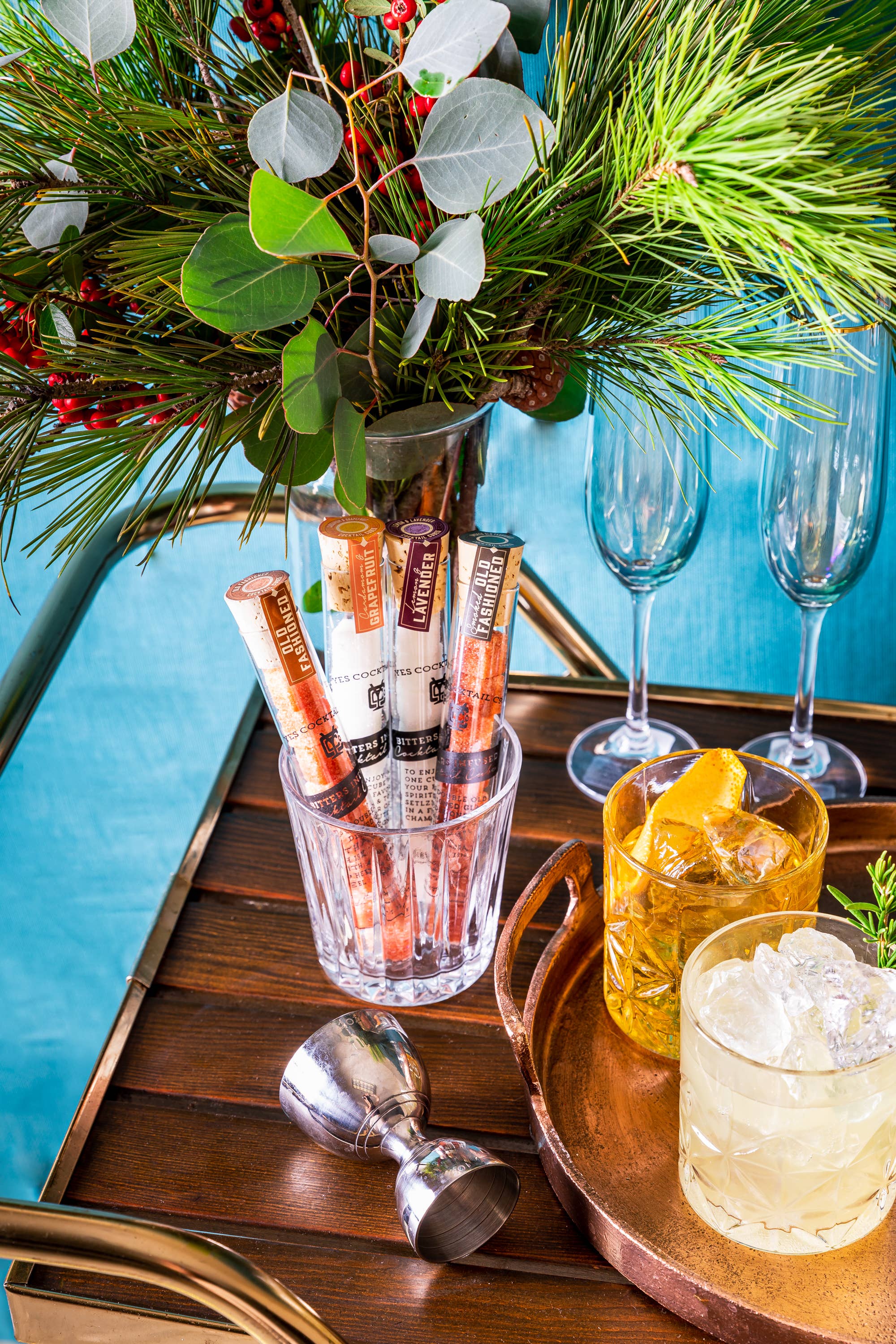 A festive bar cart is adorned with a lush arrangement of greenery and red berries. On the cart, there is a glass container holding several Ace Rivington Old Fashioned Bitters Infused Cocktail Cubes. Next to the container, there are two glasses of cocktails, one with large ice cubes and an orange garnish, and the other with crushed ice. Two champagne flutes and a metal jigger are also present on the cart.