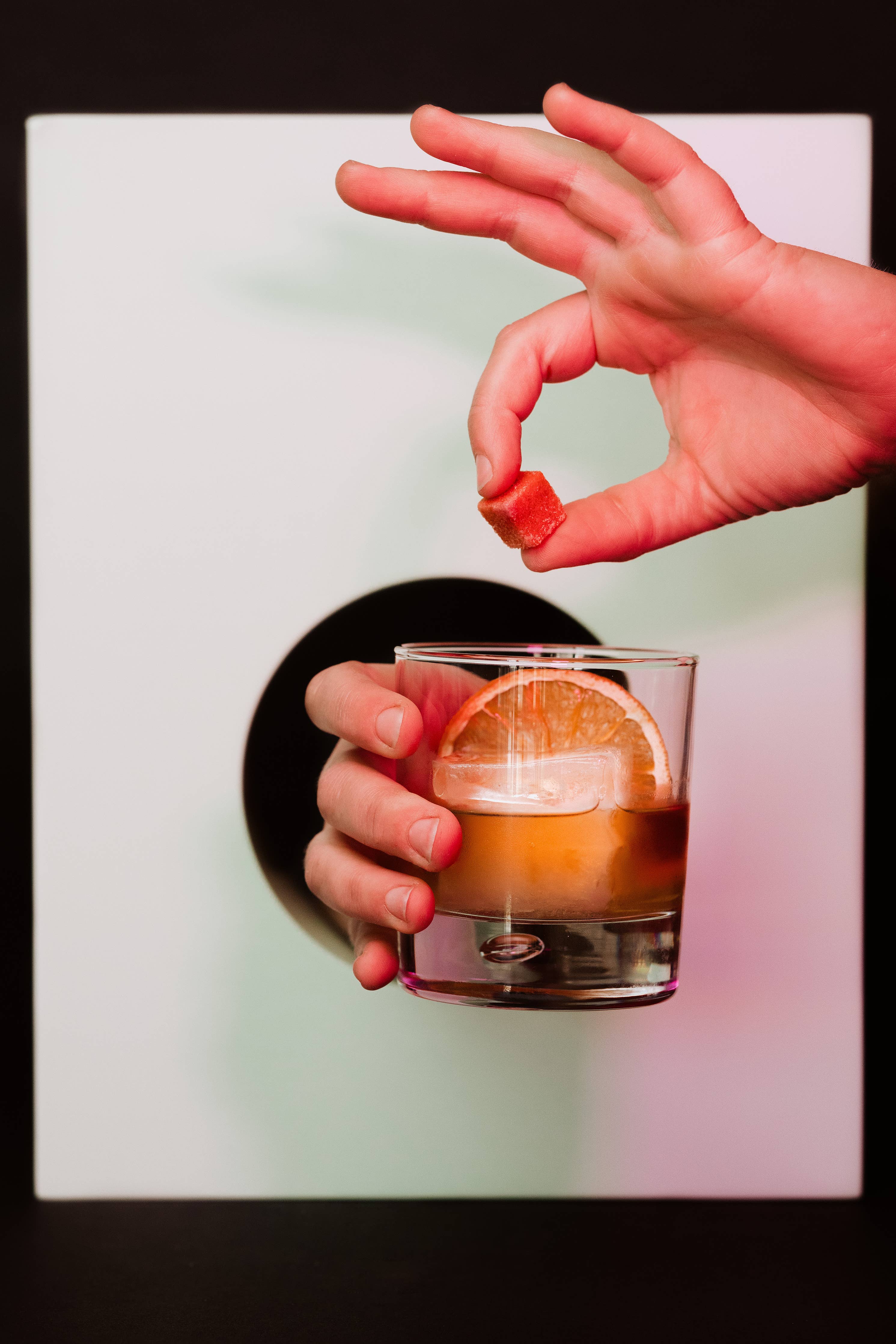 One hand holding a glass of cocktail with a slice of orange and ice cubes while another hand holds a bitters-infused cocktail cube above the glass, ready to drop it in. The background features a white surface with a black circular cutout.