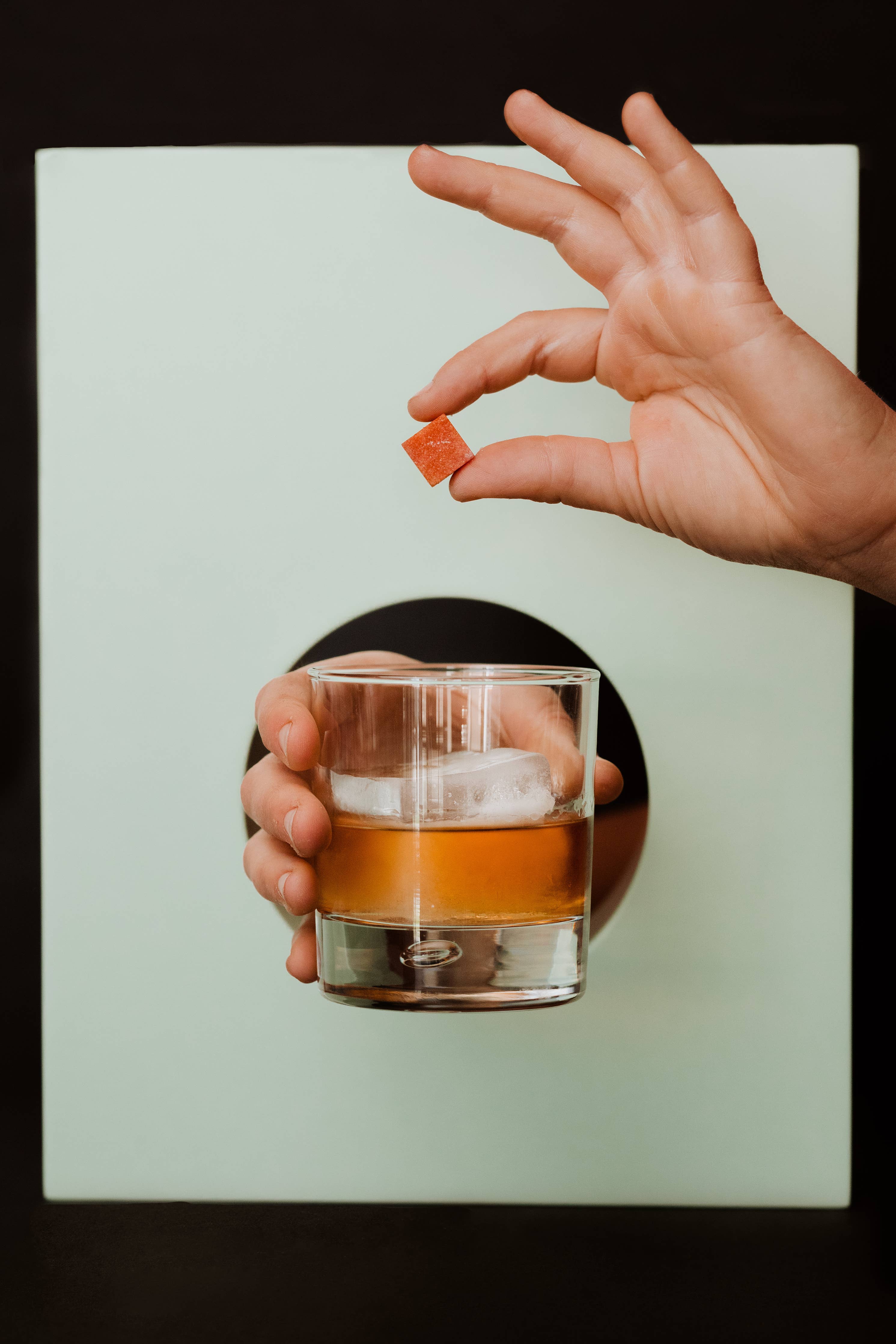 One hand is holding a glass filled with an amber-colored liquid and ice, while another hand is holding a red cocktail cube above the glass. The background features a circular cutout through which the hand holding the glass is visible. This image showcases the process of adding an Ace Rivington Old Fashioned Bitters Infused Cocktail Cube to a drink, highlighting the product's use in enhancing cocktails.