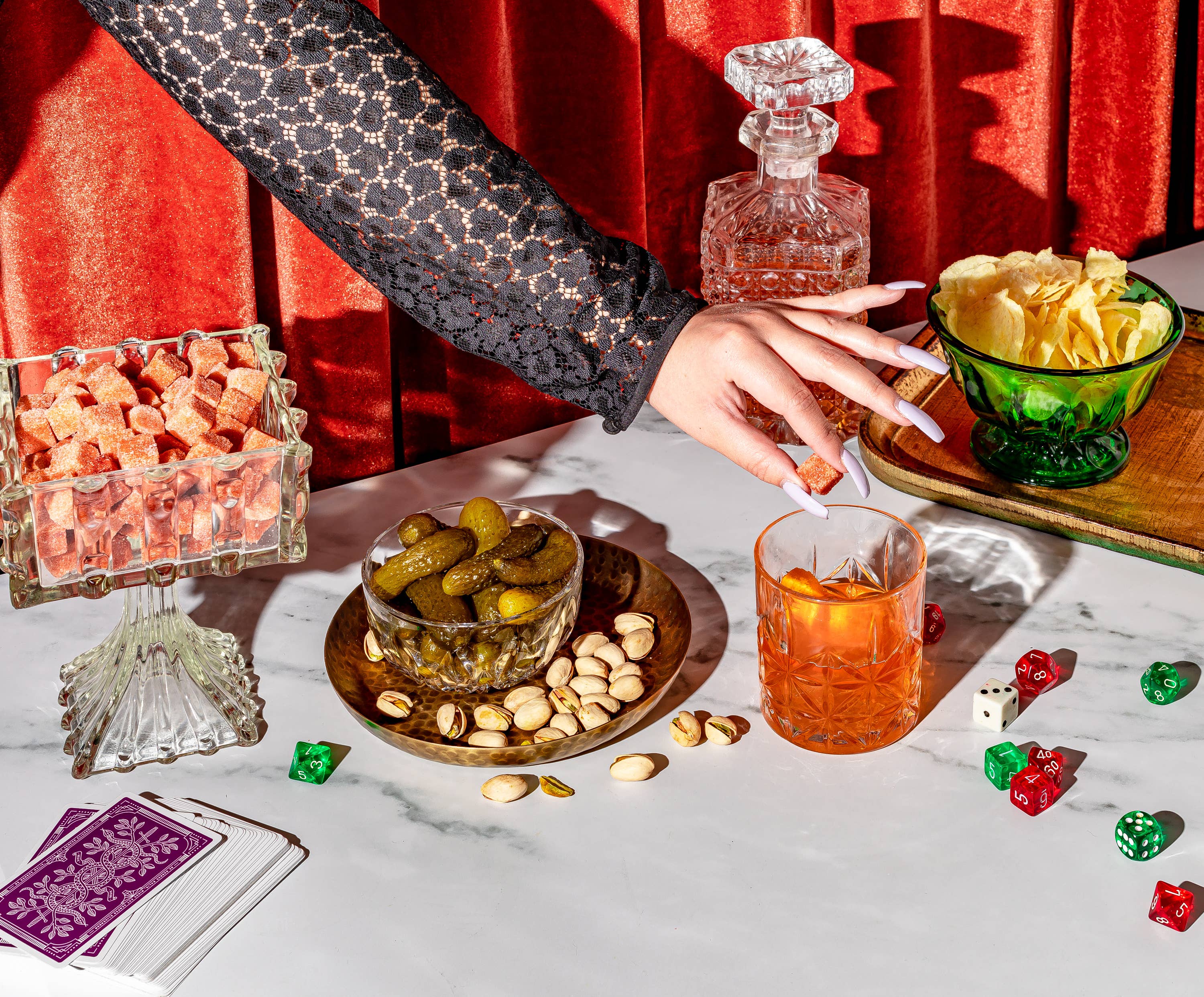 A table set with various snacks and drinks, featuring Ace Rivington Old Fashioned Bitters Infused Cocktail Cubes. A hand is reaching for a glass of cocktail with an orange slice. The table includes a bowl of pickles and pistachios, a bowl of potato chips, a glass container of red gummies, a decanter, playing cards, and dice. The background has a red curtain.