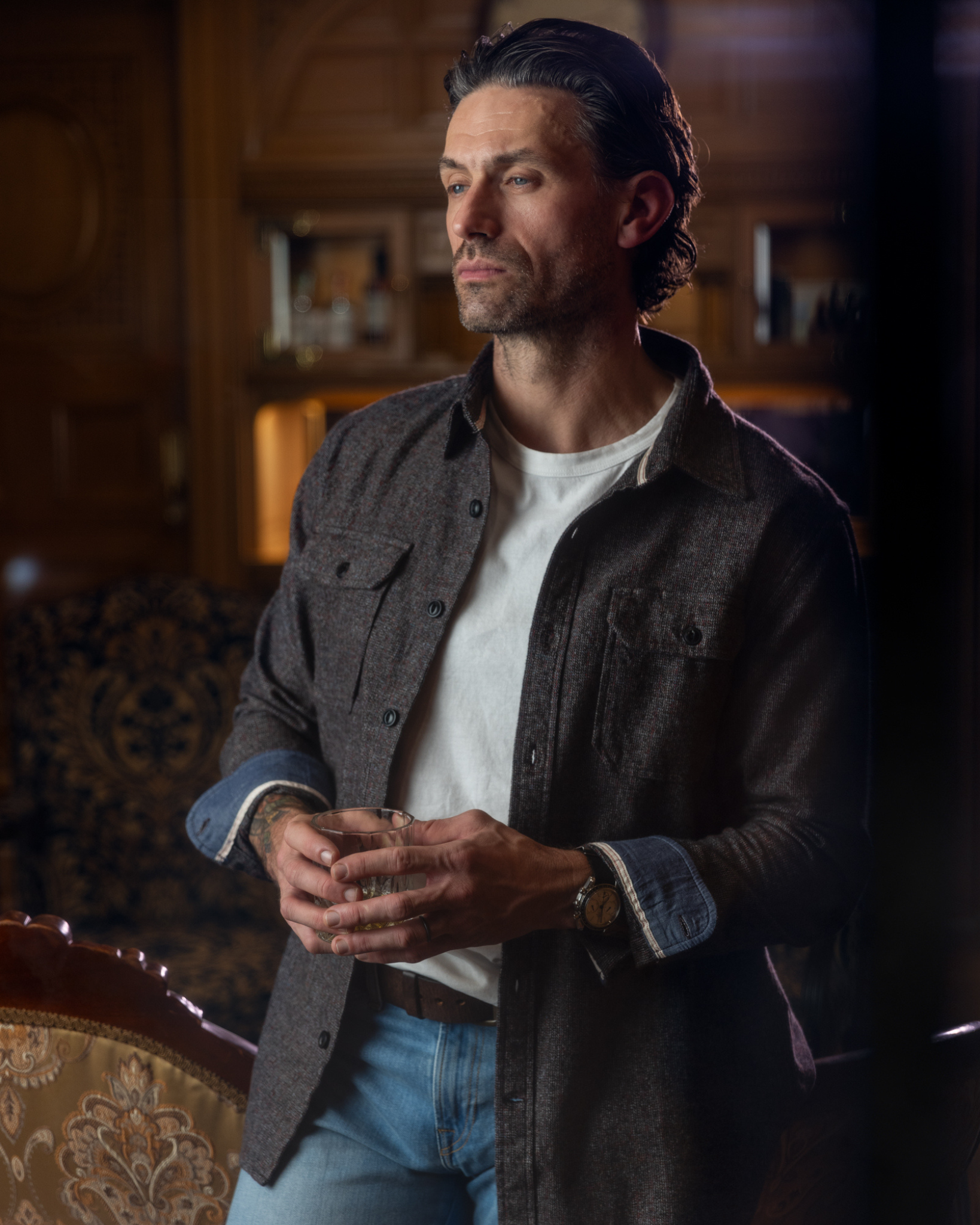 Man standing indoors wearing a white t-shirt under a dark button-up shirt with the sleeves rolled up paired with Ace Rivington - Athletic Taper Denim Jeans - Light Vintage, and a wristwatch. The background features a wooden interior with shelves and decorative elements.