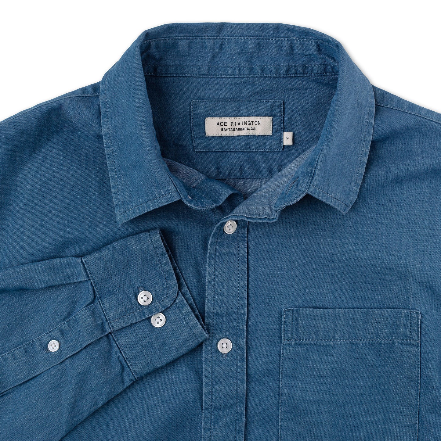 close up of cuff, collar, buttons, pocket, and tag on front of men's long sleeve tailored denim shirt in a medium wash with a single pocket and white buttons
