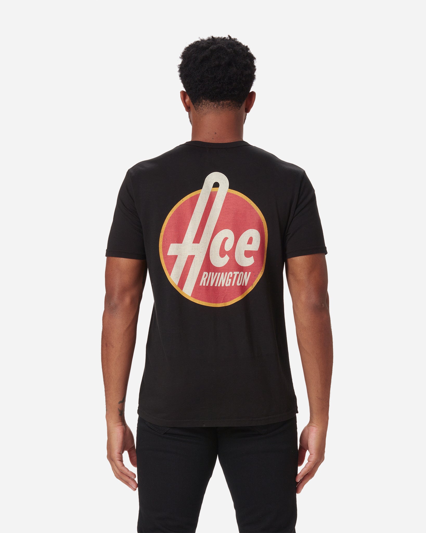 back of model wearing men's black graphic tee shirt parodying 1950's style aesthetic reading "Ace Rivington" in bold off-white text encircled by a red fill and gold rim