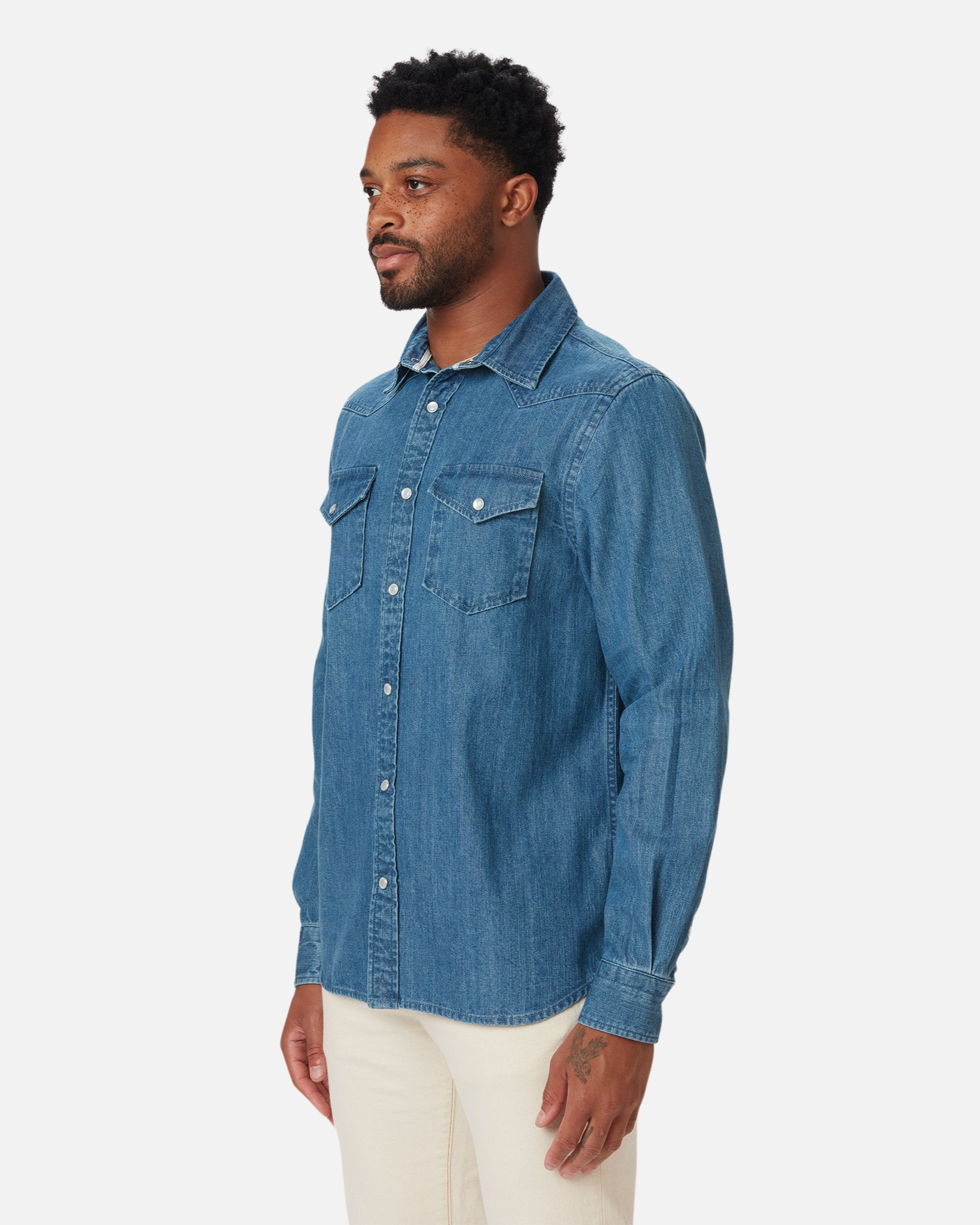 Man wearing a mid-weight denim western shirt in a medium wash by Ace Rivington features a button-down front, two chest pockets with button flaps, and long sleeves with buttoned cuffs. The shirt is paired with light-colored pants and has a classic, casual style that is both versatile and timeless, making it a relevant choice for various casual occasions.