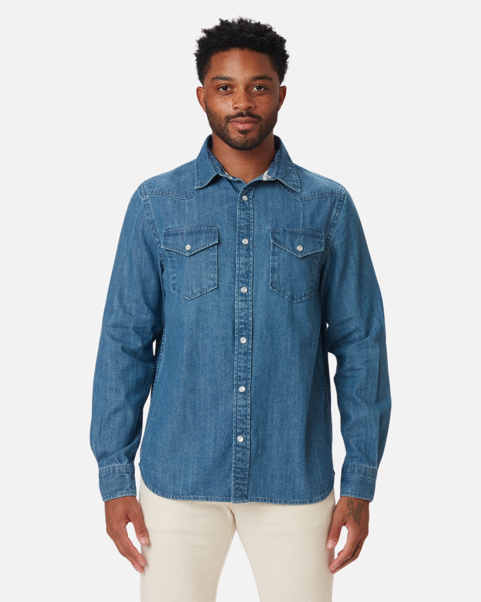 Man wearing an Ace Rivington mid-weight denim western shirt in a medium wash paired with light-colored pants. The shirt features a button-down front, two chest pockets with button flaps, and long sleeves with buttoned cuffs. The shirt has a classic Western style with a pointed collar and yoke detailing on the front and back. The background is plain white, making the shirt the focal point of the image.
