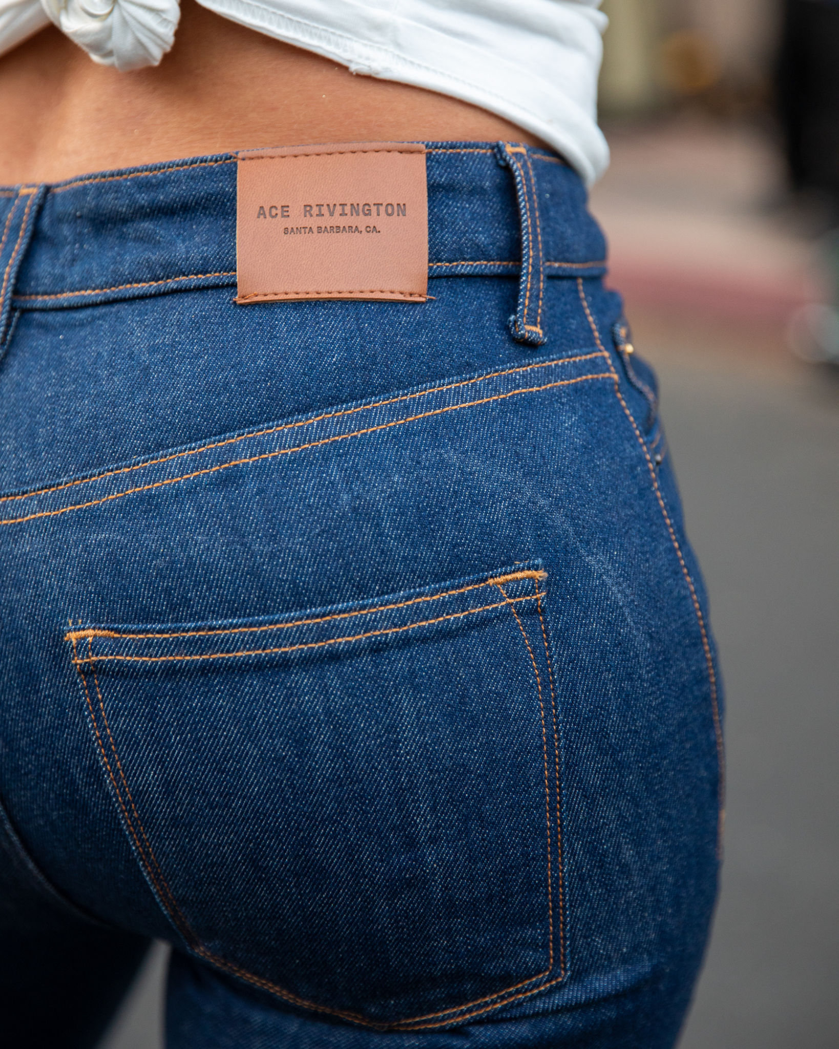 Close-up view of a woman wearing Dark Rinse - Women's Classic Straight Comfort Denim Jeans features a leather patch on the waistband with the brand name 'ACE RIVINGTON' and 'SANTA BARBARA, CA' embossed on it. The woman is also wearing a white shirt tied in a knot at the back highlighting the fit and stitching details of the jeans, including the back pocket and belt loops.