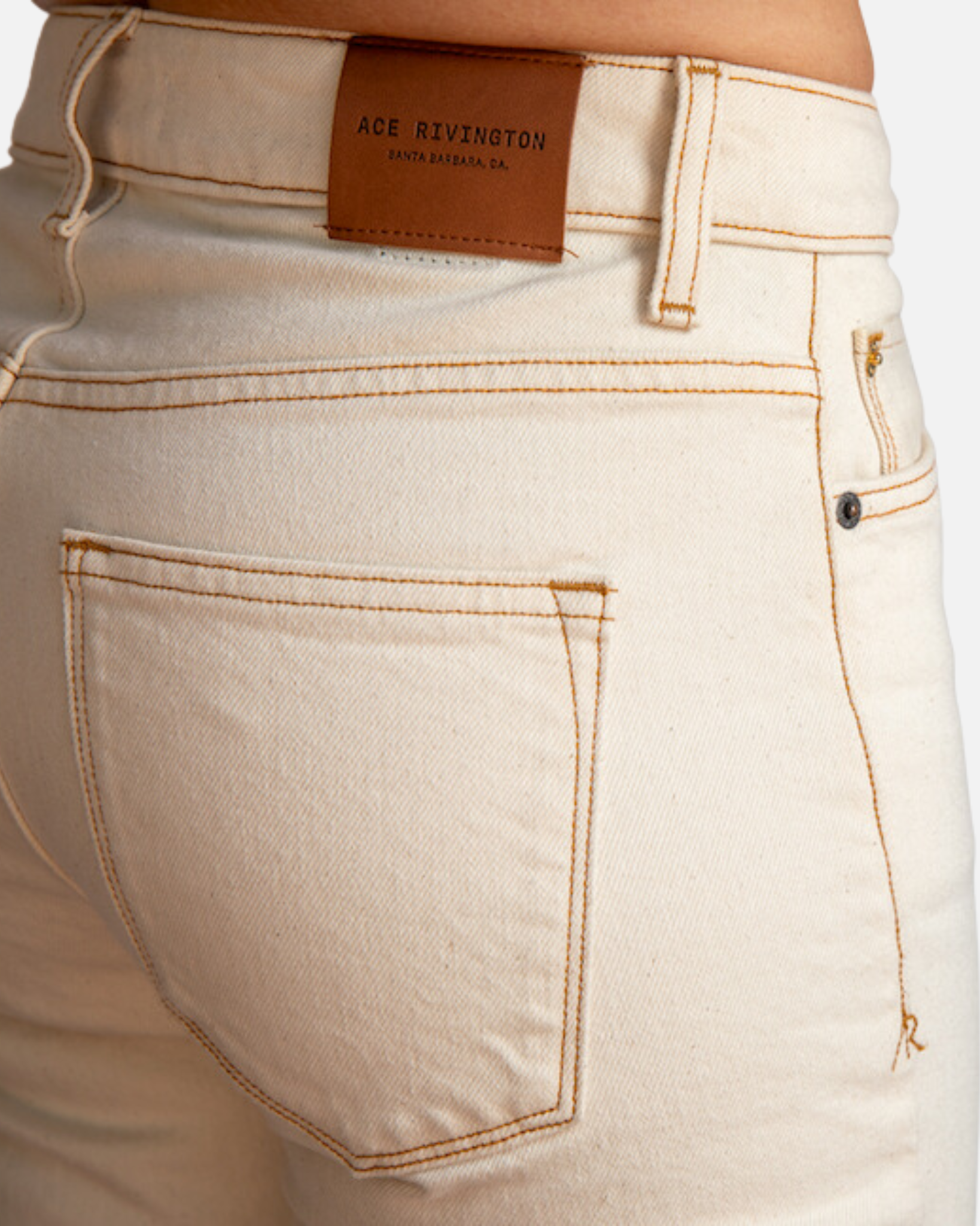 A close-up view of a woman wearing Ecru Rinse - Women's Classic Straight Comfort Denim Jeans focuses on the back pocket and waistband area, highlighting the beige color of the jeans, the brown leather patch with the brand name 'Ace Rivington' and 'Santa Barbara, CA' embossed on it, and the detailed stitching. The jeans have a classic straight fit and are made from a comfortable denim fabric.