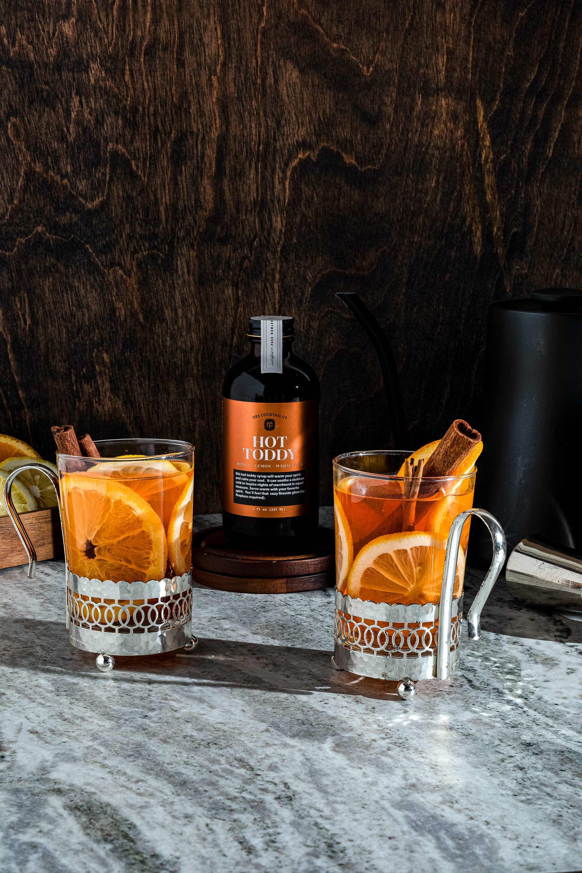 A cozy setup featuring two glass mugs filled with a hot beverage, garnished with orange slices and cinnamon sticks. Behind the mugs, there's a brown glass bottle labeled 'Hot Toddy' placed on a wooden coaster. The scene is set on a marble countertop with a dark wooden backdrop, evoking a warm and comforting atmosphere.