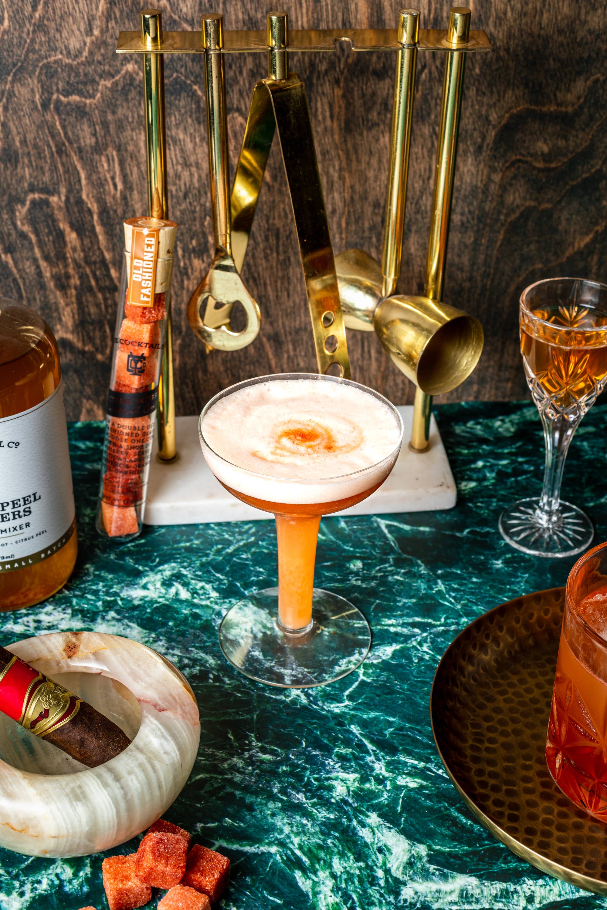 A stylish bar setup featuring Ace Rivington Old Fashioned Bitters Infused Cocktail Cubes. The scene includes a cocktail in a coupe glass with a frothy top, a bottle of cocktail mixer, a cigar in a marble ashtray, and various bar tools on a green marble surface. The cocktail cubes are visible in the foreground, adding a touch of sophistication to the arrangement.