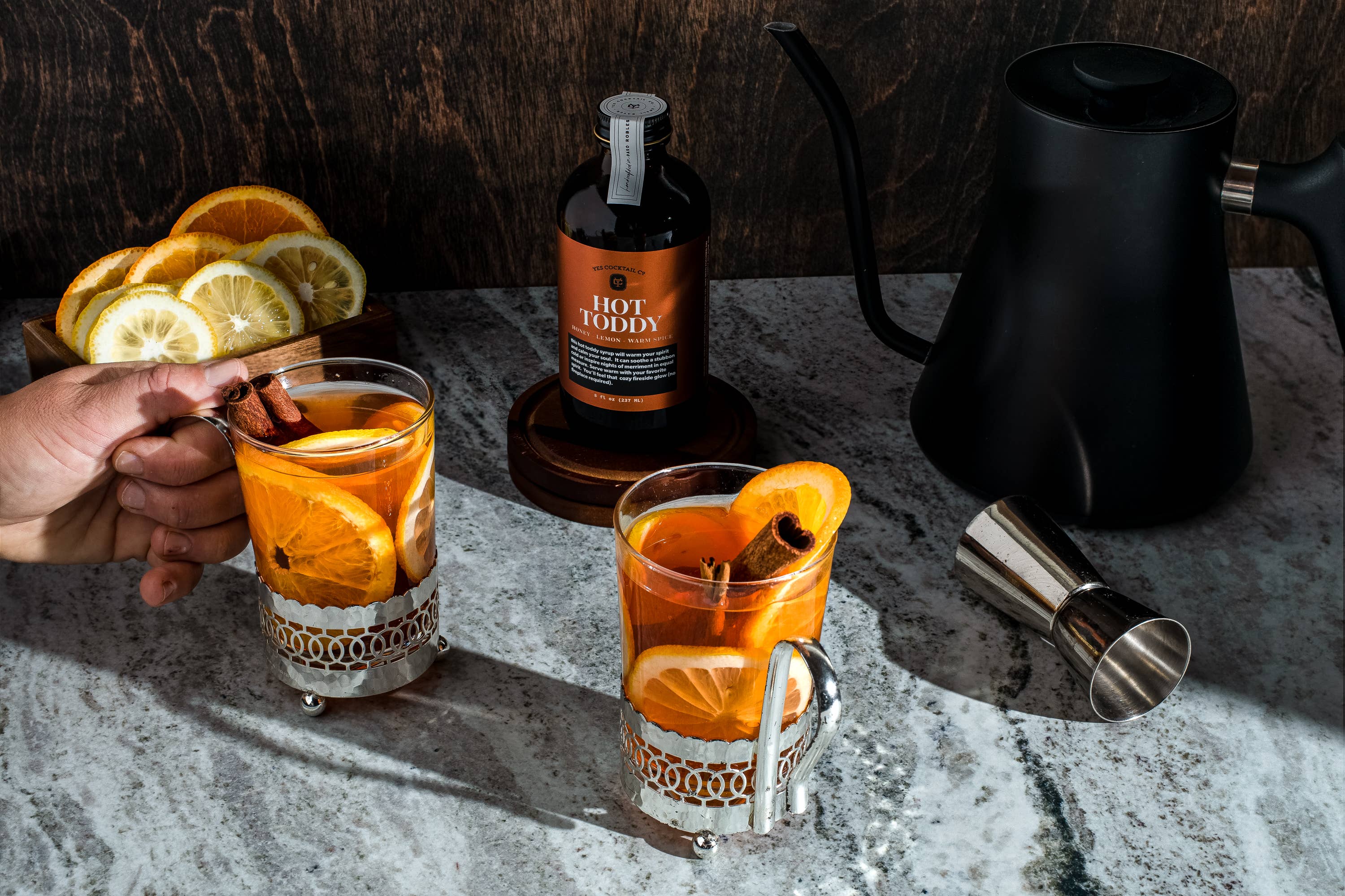 A setup for making a hot toddy with two glass mugs containing a hot beverage garnished with orange slices and cinnamon sticks. A hand is holding one of the mugs. The background features a wooden tray with sliced lemons and oranges, a bottle labeled 'Hot Toddy,' a black kettle, and a metal jigger on a marble countertop. The vibrant colors of the citrus slices and the warm tones of the drink are highlighted by the well-lit scene.