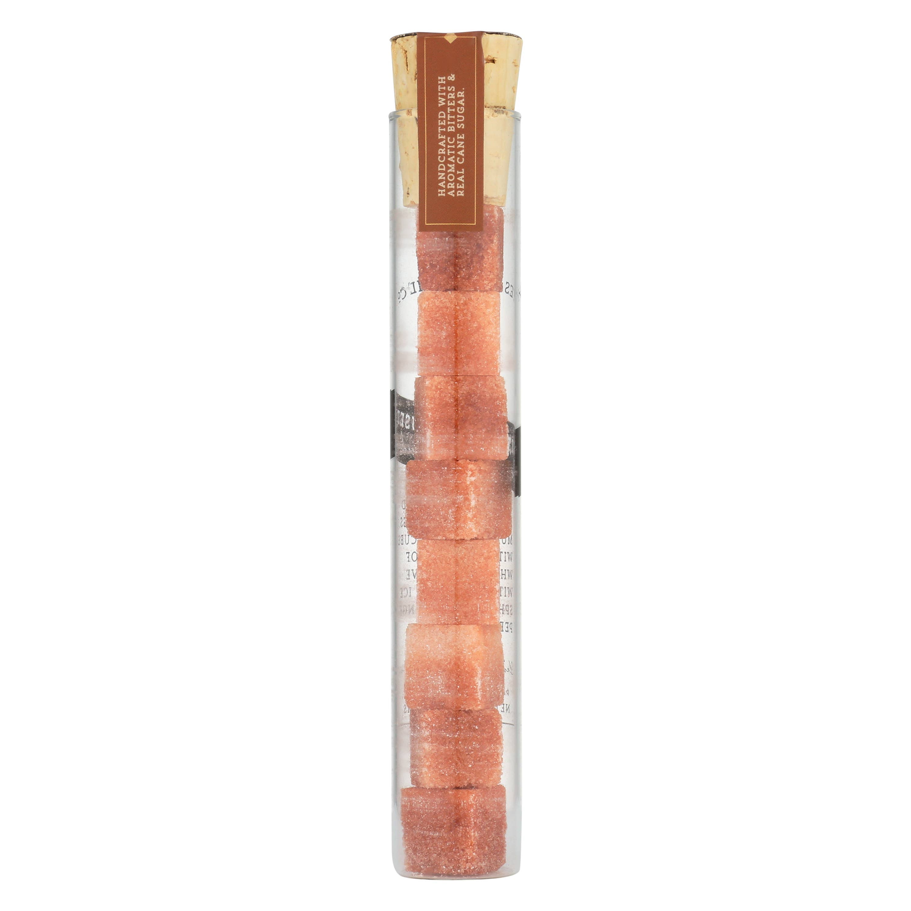 A clear cylindrical container with a cork stopper, holding a stack of pinkish-orange cocktail cubes. The label on the container reads 'Handcrafted with Aromatic Bitters & Real Cane Sugar'