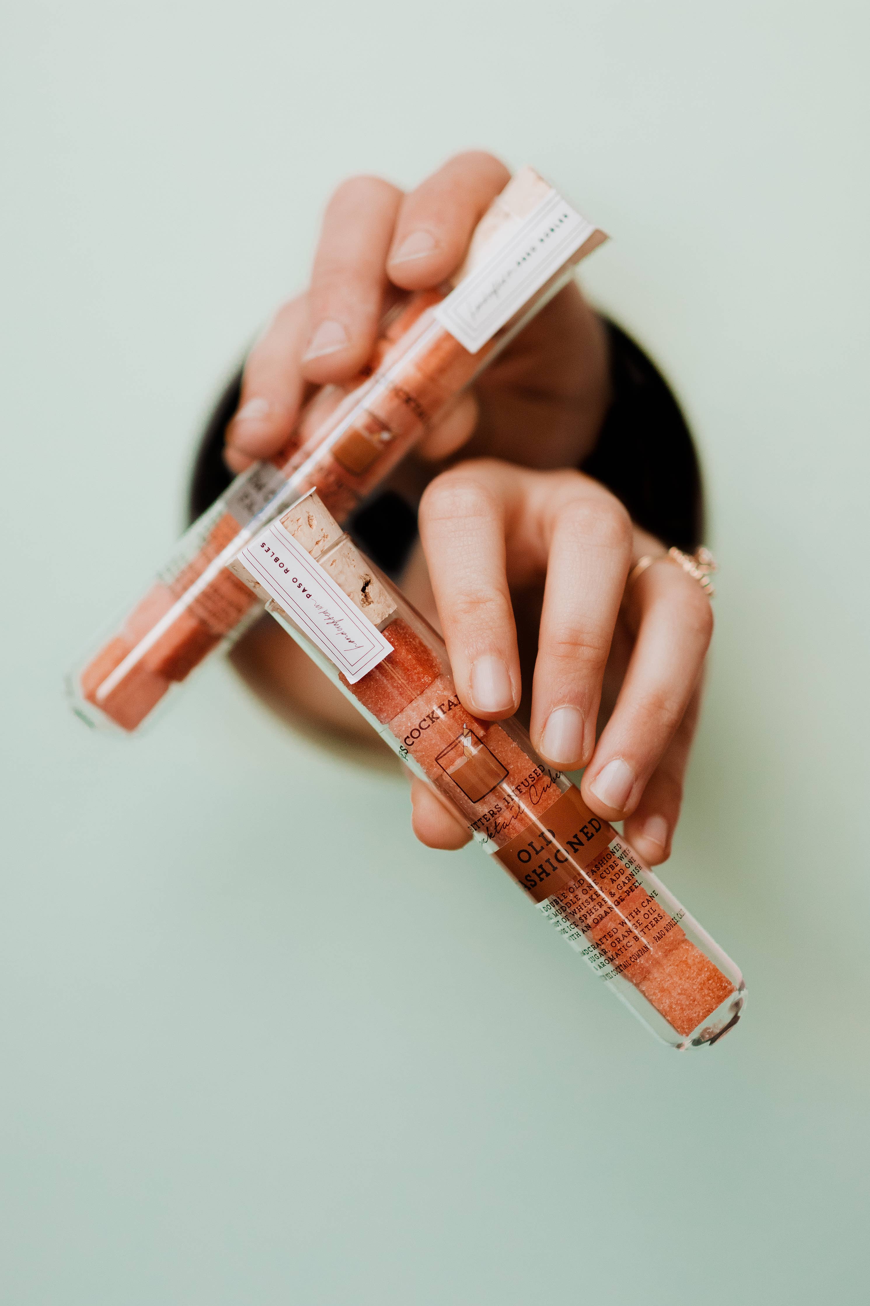 A pair of hands holding two transparent tubes containing Ace Rivington Old Fashioned Bitters Infused Cocktail Cubes against a light background. The tubes have labels with text and the cubes inside are orange in color.