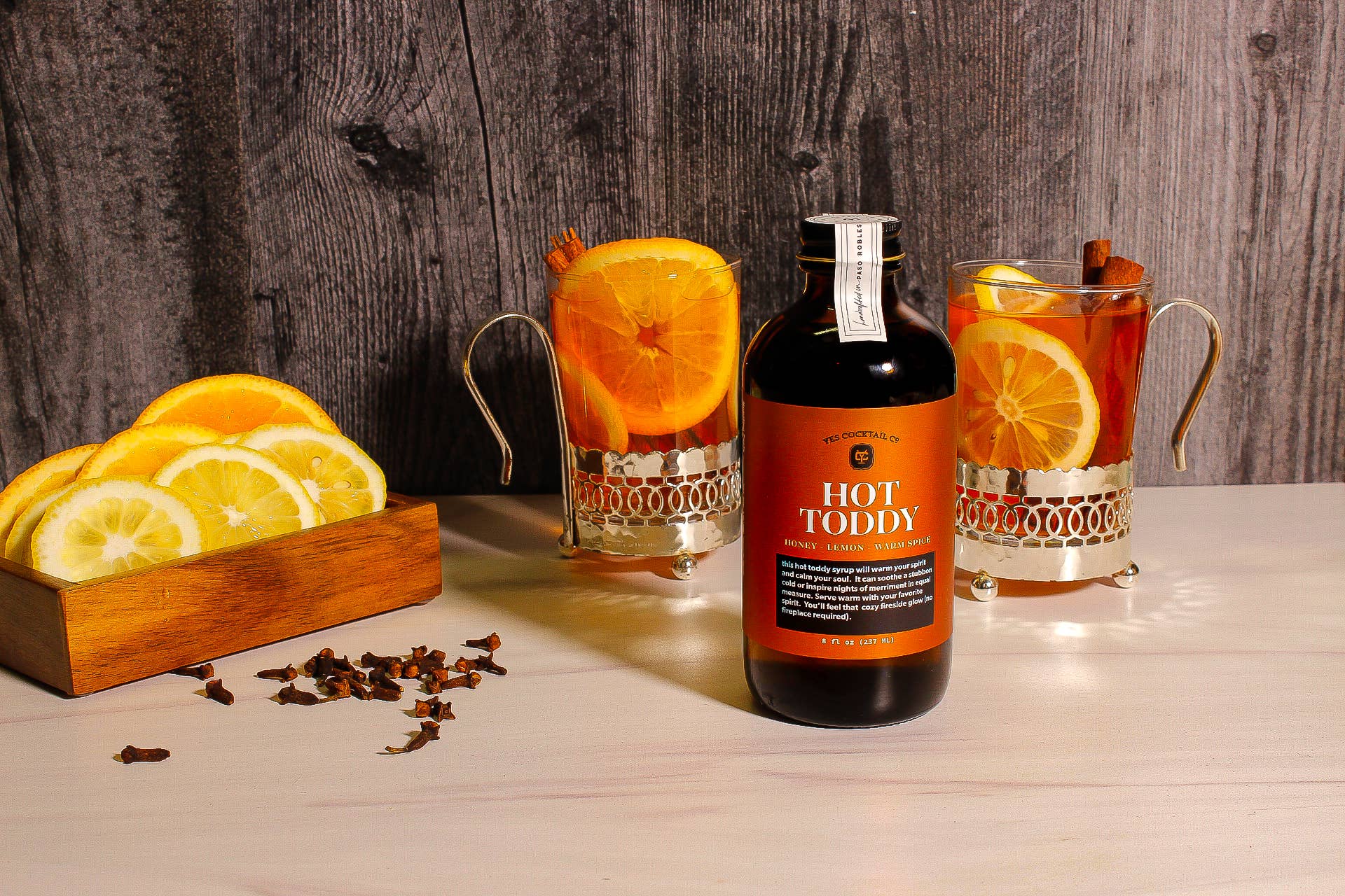 A bottle of Ace Rivington Hot Toddy Syrup is placed on a wooden table with a label that reads 'Hot Toddy, Honey, Lemon, Ginger.' Two glass mugs next to the bottle contain a beverage garnished with orange slices and cinnamon sticks. There is also a wooden container with lemon slices and some scattered cloves on the table. The background features a wooden surface.