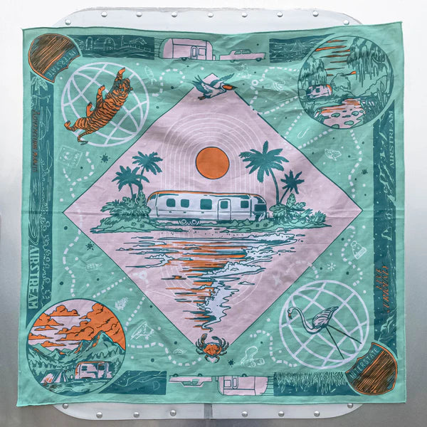 The Ace Rivington Bandits Bandana - Live Curiously features a pastel pink and green background with an Airstream trailer by a body of water and palm trees. It includes circular illustrations of a tiger, a flamingo, a mountain landscape, and a river trailer. Small border illustrations of trailers and nature scenes accompany the text 'AIRSTREAM' and 'LIVE CURIOUSLY,' evoking a spirit of adventure.