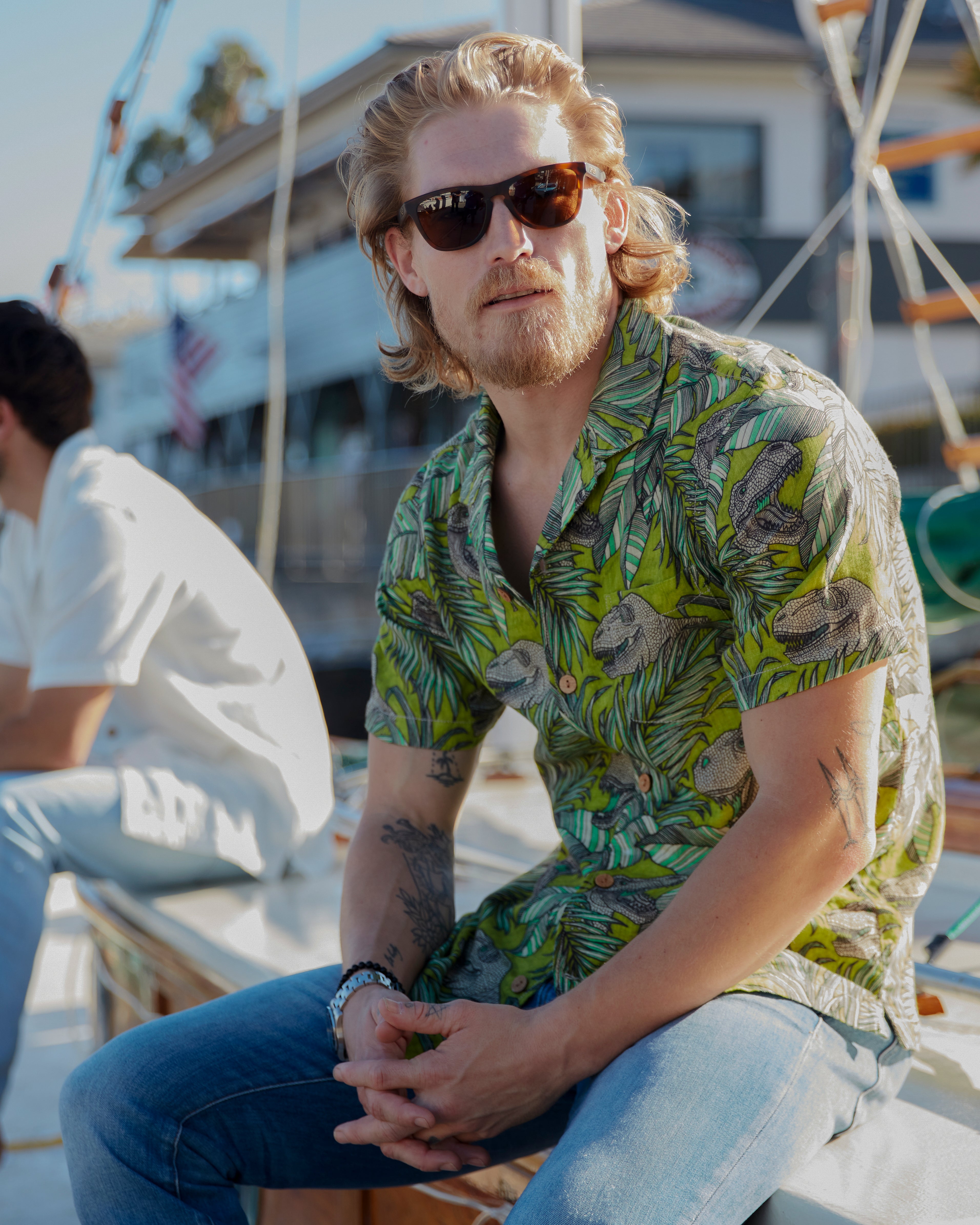 Side of model with a front and leftward gaze sitting on a boat in the Santa Barbara Harbor wearing sunglasses and Ace Rivington "Man Eater" green and brown floral-themed camp shirt with leaf and dinosaur design with left breast-pocket made with doubles-gauze soft-textured cotton fabric and brown wooden buttons and medium vintage Ace Rivington blue denim jeans