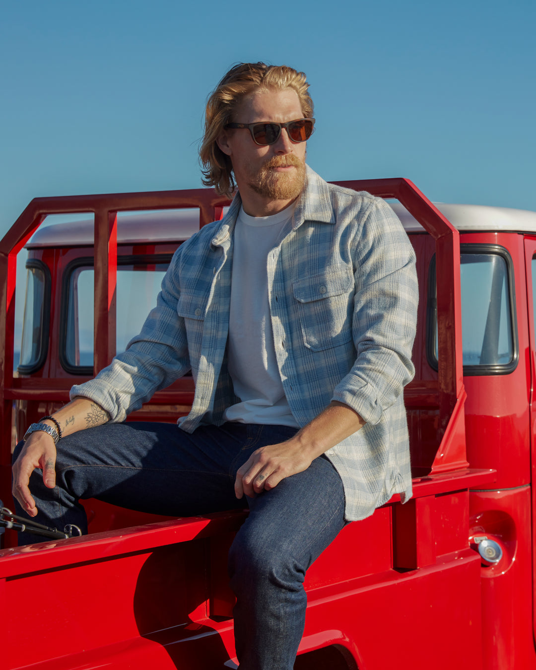 Lifestyle-oriented model sitting on the bed of a cherry red Volkswagen Van pickup conversion with a slight leftward gaze wearing sunglasses, f Ace Rivington soft-brushed flannel shirt with pearl buttons and off-white and light blue and black checkered grid plaid design with pearl buttons and double breast pockets with buttoned enclosure, and pair of Ace Rivington dark clean denim jeans with dark, rich indigo color and no fabric wear
