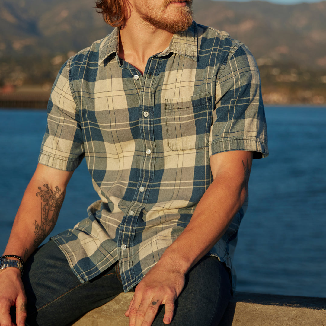 Lifestyle-oriented model sitting with a leftward gaze facing away from the ocean and mountains of Santa Barbara wearing Ace Rivington collared short-sleeved tailored 100% cotton shirt with real indigo slight blue and beige plaid patterning and left breast pocket and a pair of Ace Rivington dirty vintage denim jeans with slight stylistic fabric wear