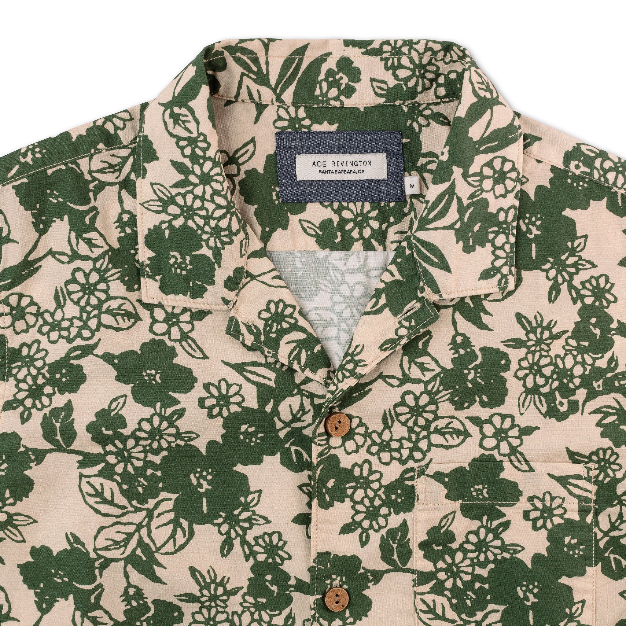 close up of tag, buttons and pocket on front of men's short sleeve button up camp shirt with hawaiian style design with beige background and green flowers with wooden buttons