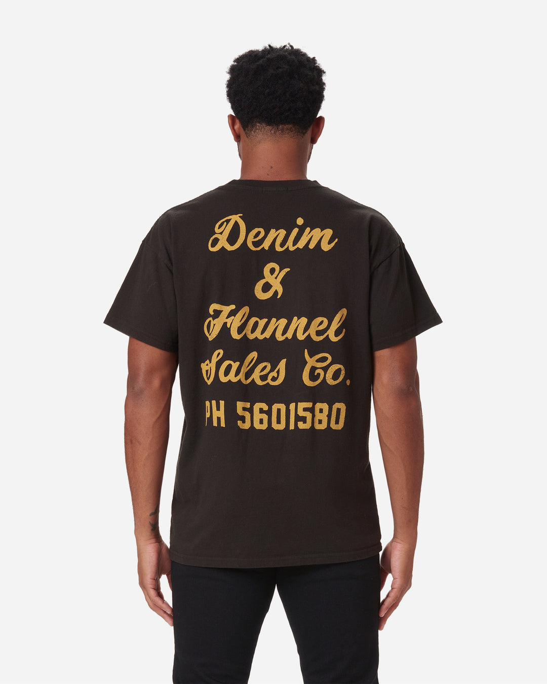 back of model wearing men's black graphic tee with gold lettering reading "denim & flannel sales co. ph 5601580"