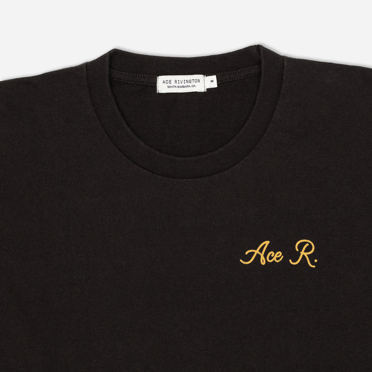 zoom in of lettering on front of men's black graphic tee with gold cursive lettering reading "Ace R." over the left breast area and Ace Rivington brand logo tag