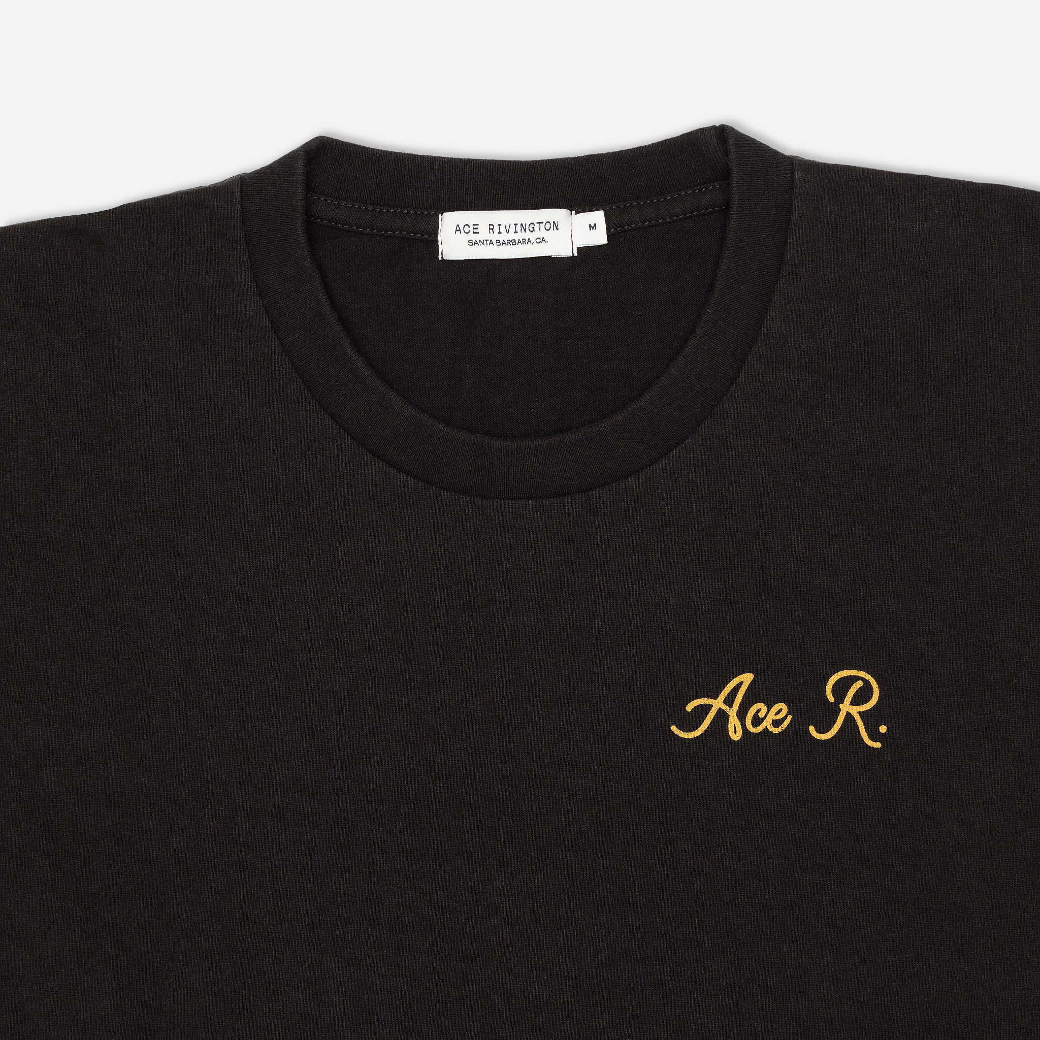 zoom in of lettering on front of men's black graphic tee with gold cursive lettering reading "Ace R." over the left breast area and Ace Rivington brand logo tag