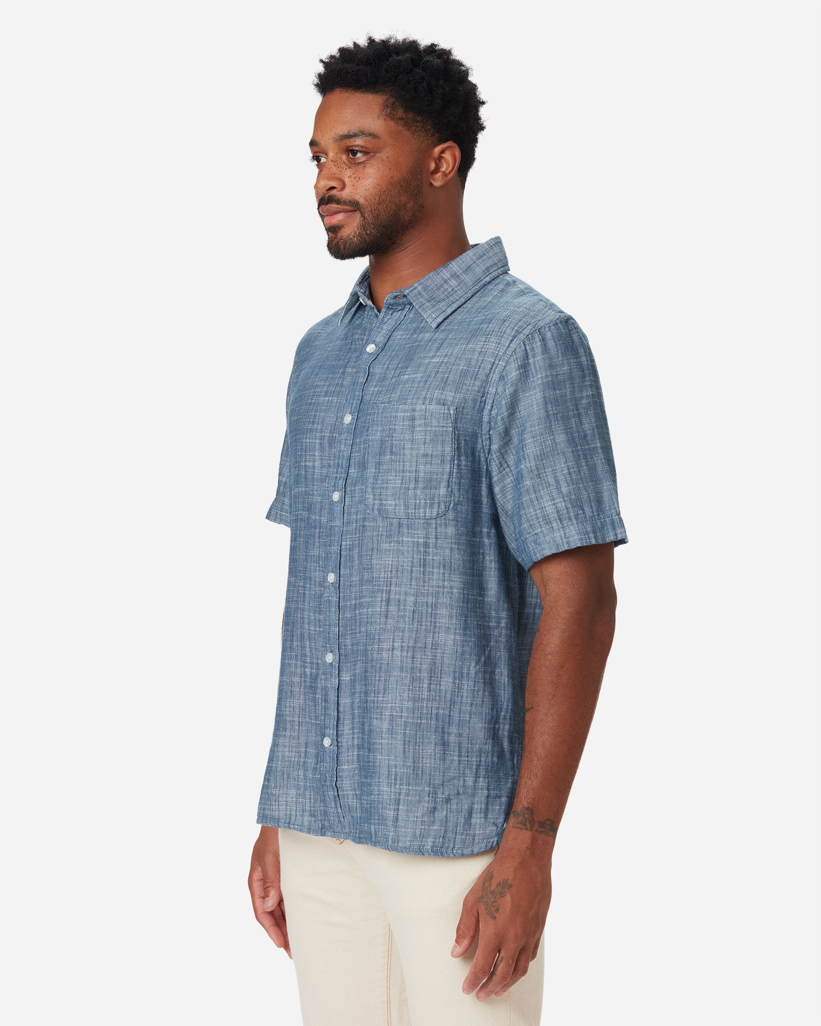 Model with front and rightward gaze wearing Ace Rivington short-sleeved indigo slub soft textured-cotton shirt with linen hand feel, pearl buttons, left-breast pocket, collar, and off white interior and pair of Ace Rivington ecru color denim jeans