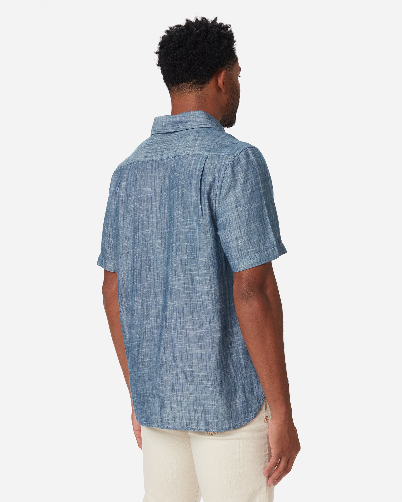 back of model facing away with a slight rightward gaze wearing Ace Rivington short-sleeved indigo slub soft textured-cotton shirt with linen hand feel, pearl buttons, left-breast pocket, collar, and off white interior and a pair of Ace Rivington ecru color denim jeans