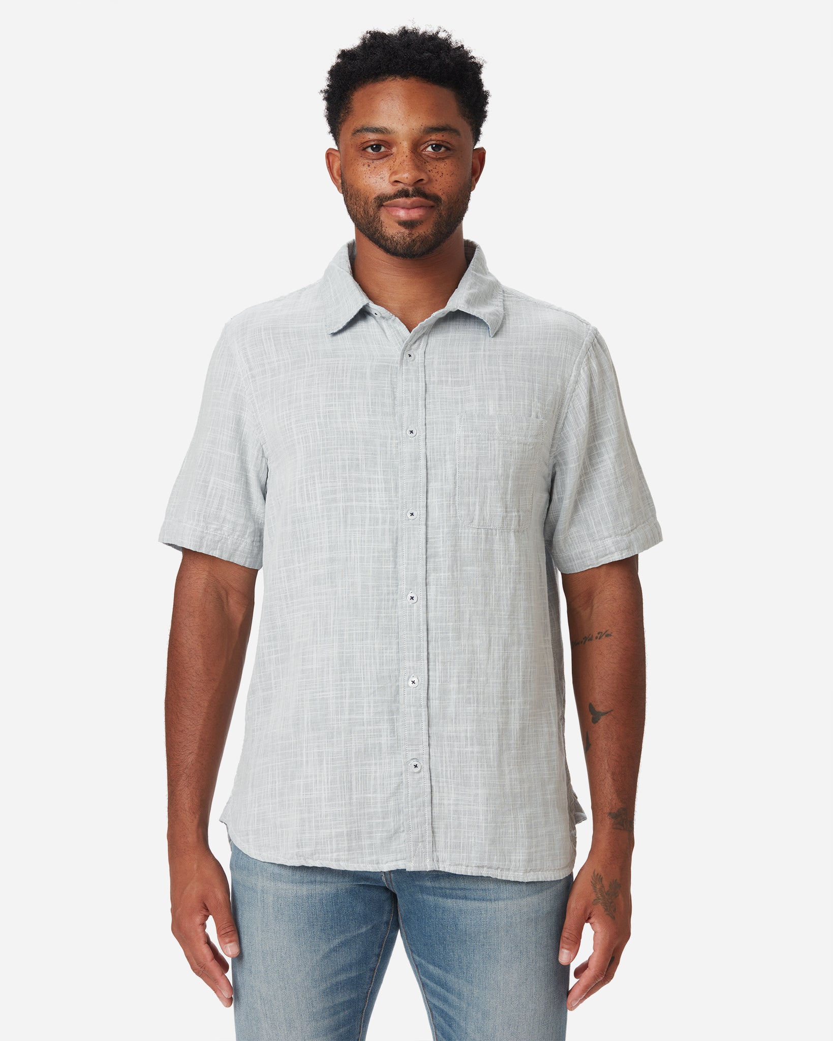Model with direct frontward gaze wearing Ace Rivington collared grey short-sleeved double gauze soft-textured cotton shirt with indigo blue interior, pearl buttons, and left-breast pocket and a pair of Ace Rivington medium vintage medium light blue denim jeans