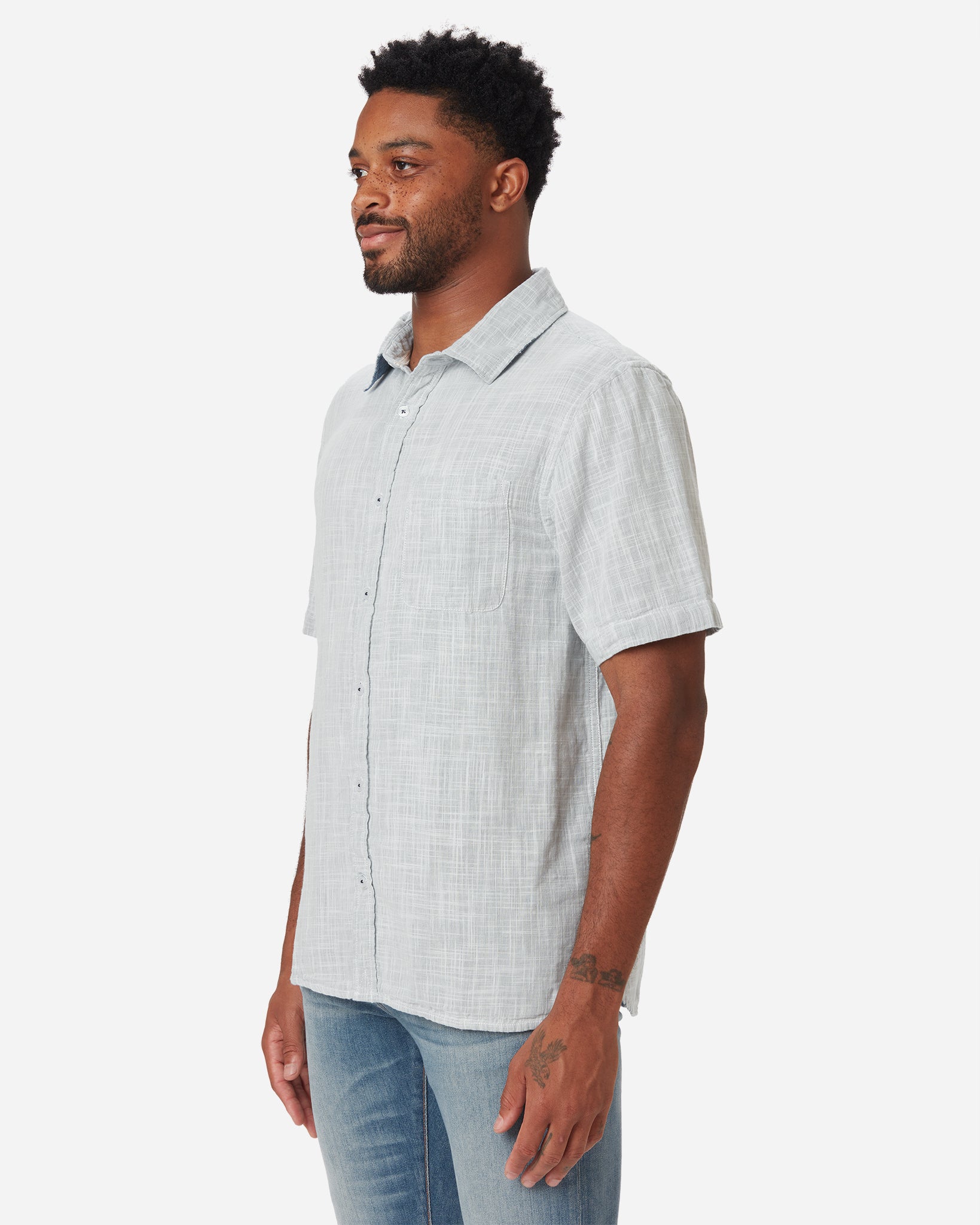 Model with a forward and rightward gaze wearing Ace Rivington collared grey short-sleeved double gauze soft-textured cotton shirt with indigo blue interior, pearl buttons, and left-breast pocket and a pair of Ace Rivington medium vintage medium light blue denim jeans 
