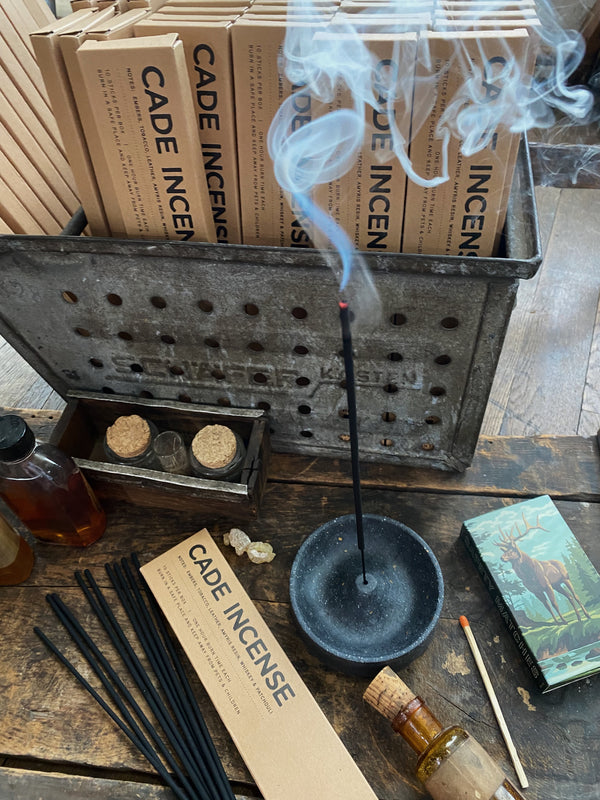 A display of Cade Incense products shows a metal container filled with boxes labeled 'CADE INCENSE.' In front of the container, there is a lit incense stick in a holder, emitting smoke. Various items are arranged on the wooden table, including a bottle with a cork, a box with small glass vials, a matchbox with a deer illustration, and several loose incense sticks. The scene has a rustic and natural aesthetic, highlighting the incense product.
