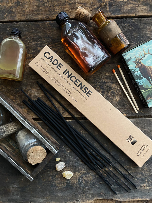 A rustic wooden table displays a box labeled 'CADE INCENSE' with black incense sticks, two glass bottles with corks, a small wooden crate with jars, a book with a deer illustration, and a few matches. The Cade Incense box highlights notes of cade, birch tar, tobacco, and other scents. The scene evokes a vintage, artisanal atmosphere.