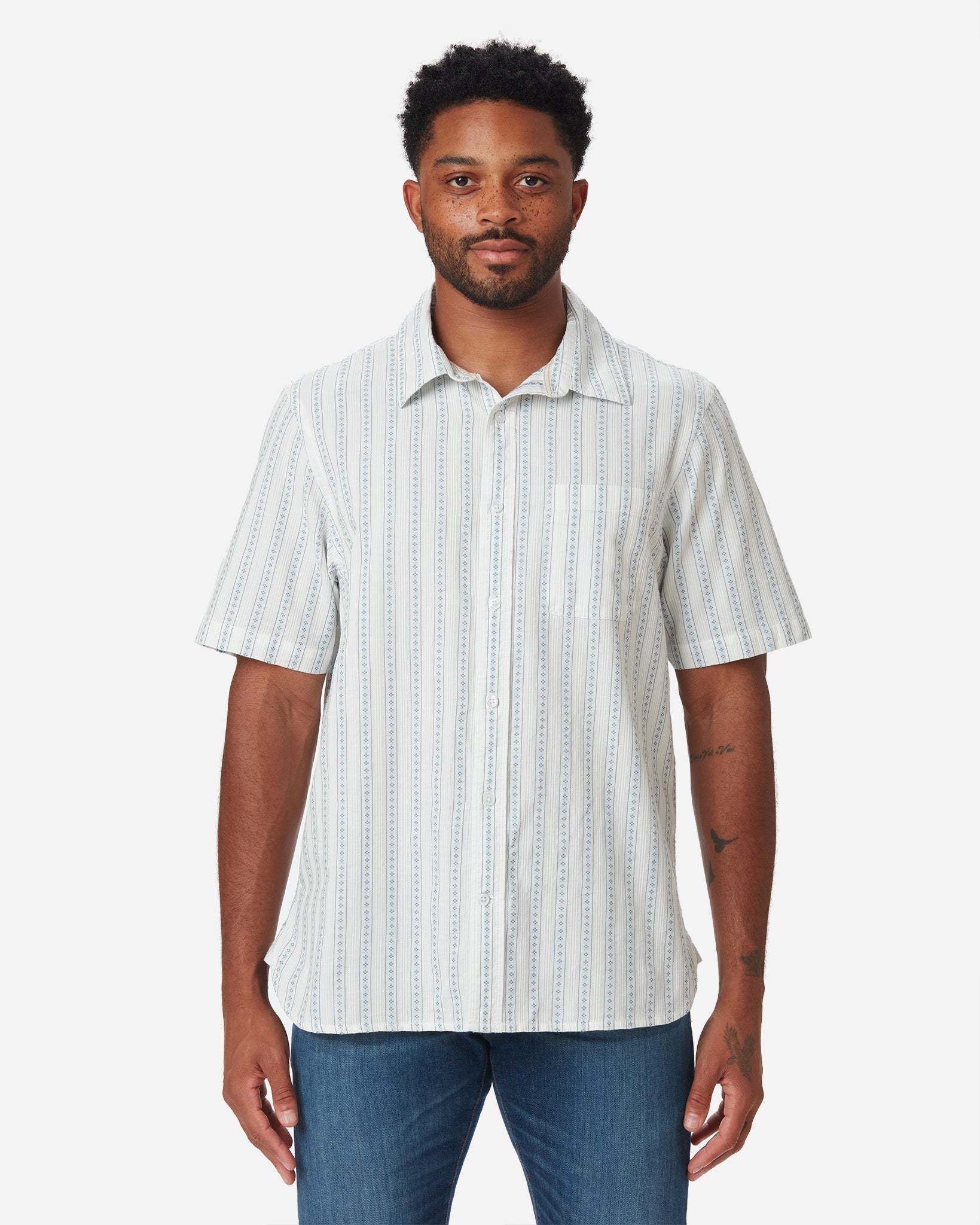 Model with directly forward gaze wearing Ace rivington light-weight tailored shirt in off-white with light blue vertical stripes and dark blue diamond pattern within each stripe with pearl buttons and left-breast pocket and pair of Ace Rivington dirty vintage blue denim jeans with slight stylistic wear