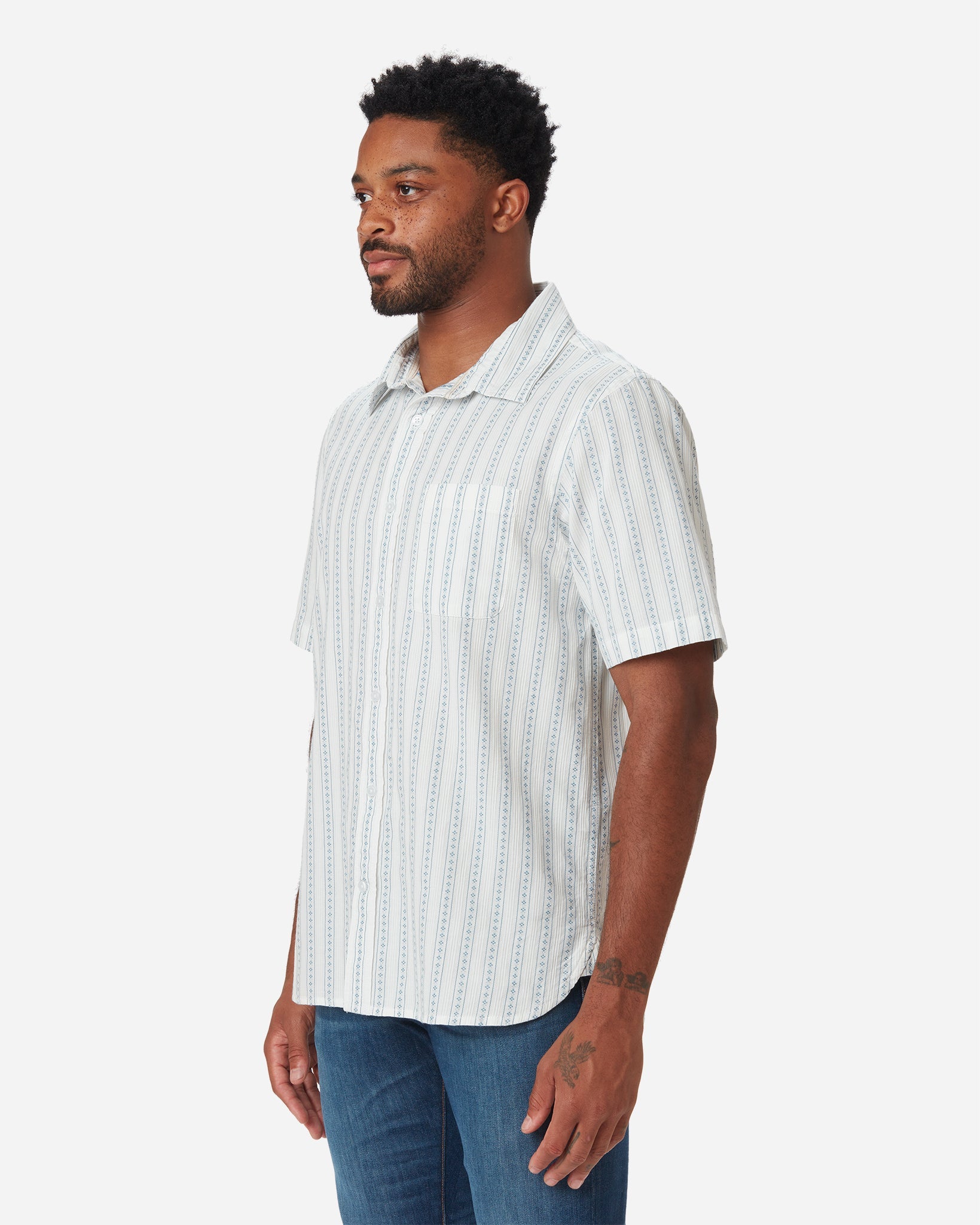 Model with a front and rightward gaze wearing Ace rivington light-weight tailored shirt in off-white with light blue vertical stripes and dark blue diamond pattern within each stripe with pearl buttons and left-breast pocket and pair of Ace Rivington dirty vintage blue denim jeans with slight stylistic wear