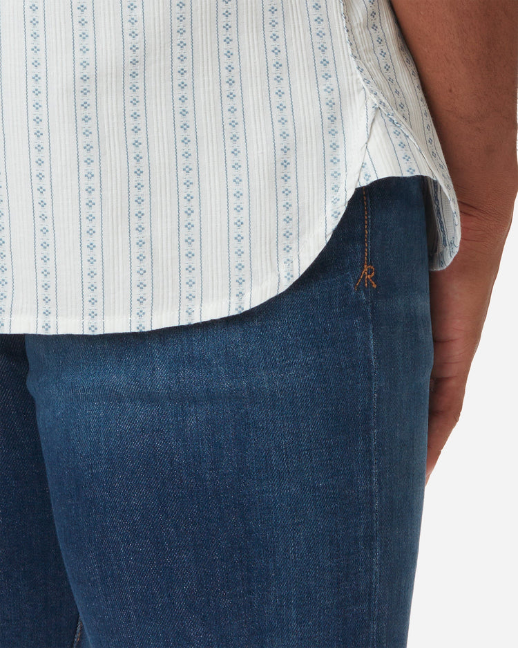 Zoomed in detail shot of the hem and top of the jeans with signature "AR" on model Model facing away with slightly rightward gaze wearing Ace rivington light-weight tailored shirt in off-white with light blue vertical stripes and dark blue diamond pattern within each stripe with pearl buttons and left-breast pocket and pair of Ace Rivington dirty vintage blue denim jeans with slight stylistic wear