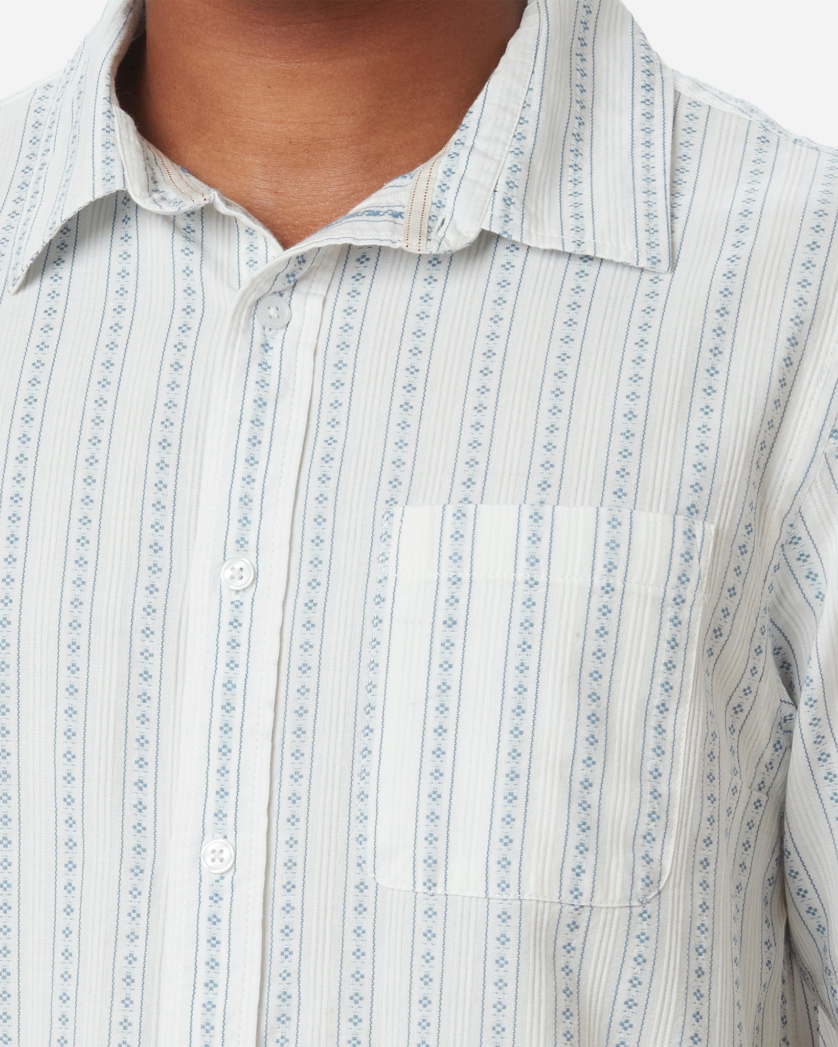 Zoomed-in detail of neck, buttons, breast pocket, and collar on model Model facing away with slightly rightward gaze wearing Ace rivington light-weight tailored shirt in off-white with light blue vertical stripes and dark blue diamond pattern within each stripe with pearl buttons and left-breast pocket and pair 