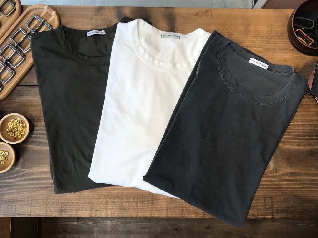 assorted plain men's t shirts in black gray and off white on a rustic wood grain table with other clothing materials