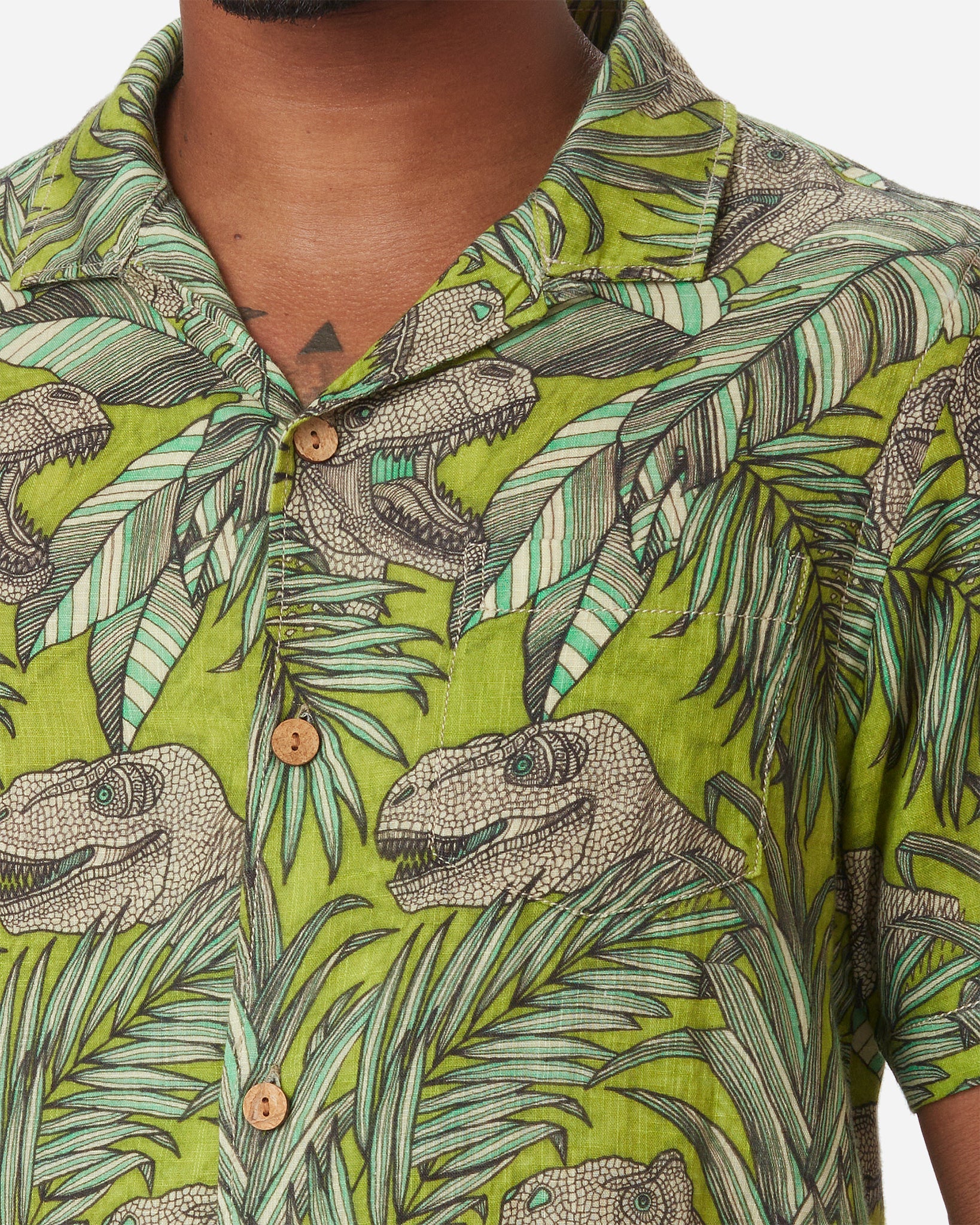 Zoomed-in detail of neck, left-breast pocket, and buttons on model with a front and slightly rightward gaze wearing Ace Rivington "Man Eater" green and brown floral-themed camp shirt with leaf and dinosaur design with left breast-pocket made with doubles-gauze soft-textured cotton fabric 