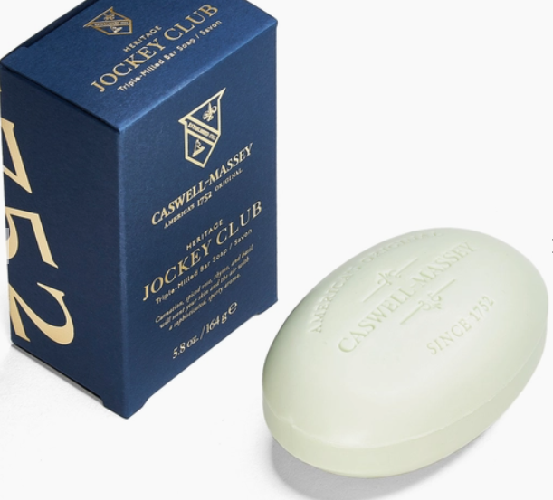 Ace Rivington Caswell-Massey Heritage Jockey Club soap is displayed next to its blue packaging box features gold text and a crest, stating 'Caswell-Massey American 1752 Original' and 'Heritage Jockey Club Triple-Milled Bar Soap.' The soap bar itself is oval-shaped and light green, embossed with 'Caswell-Massey Since 1752' highlighting the soap's elegant packaging and premium quality.