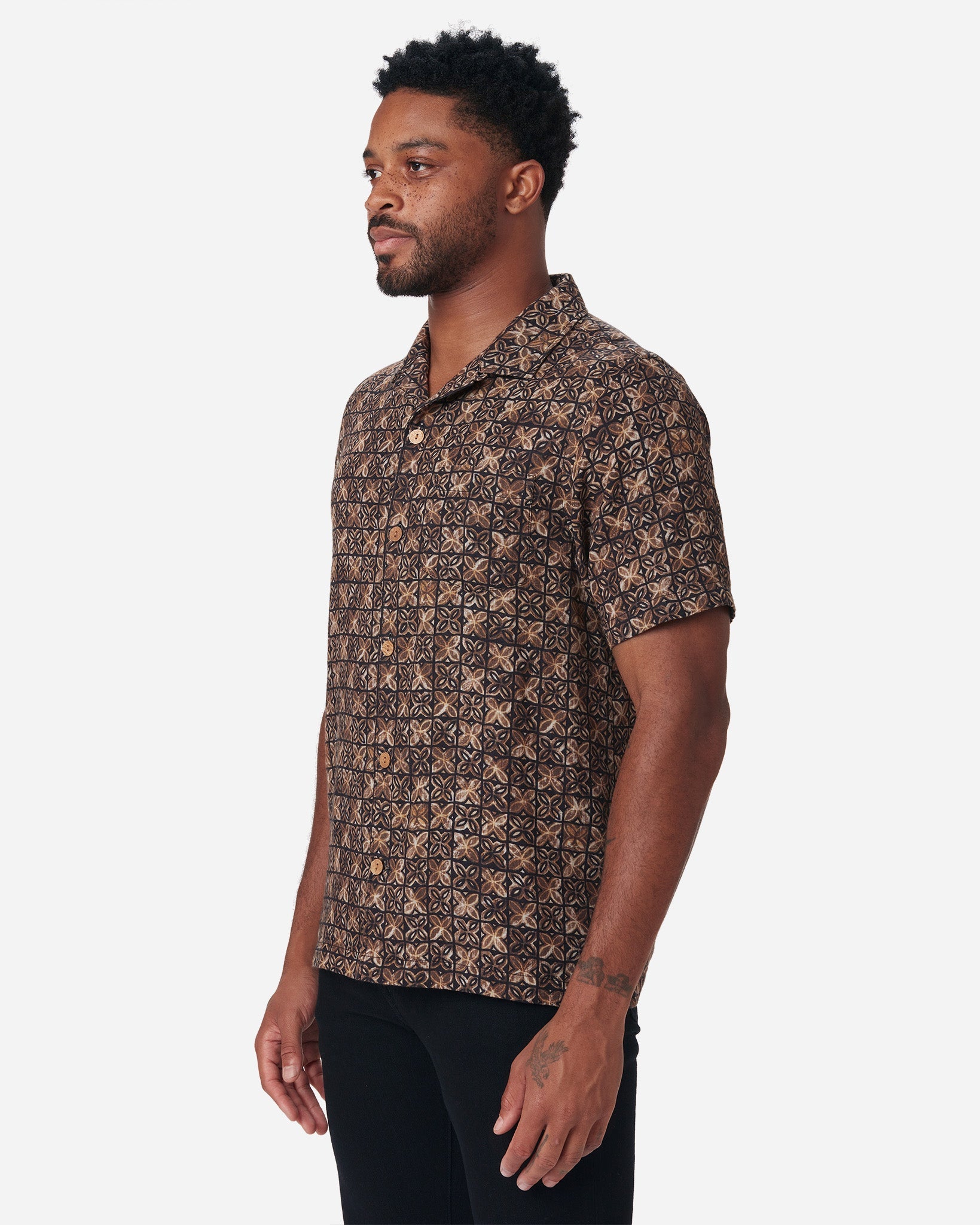 Model with a front and rightward gaze wearing Ace Rivington "Vintage Tile" gold, brown, and black exclusive design camp-collar shirt with coconut buttons and doubles-gauze soft-textured cotton fabric and a pair of black Ace Rivington denim jeans