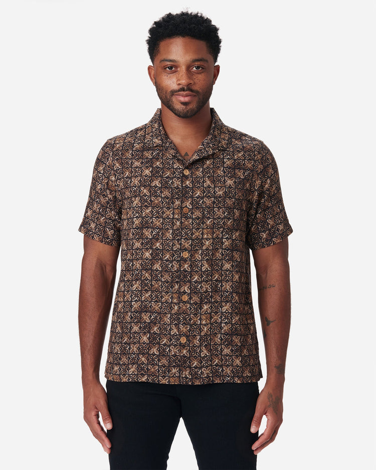 Directly frontward gaze of model wearing Ace Rivington "Vintage Tile" gold, brown, and black exclusive design camp-collar shirt with coconut buttons and doubles-gauze soft-textured cotton fabric and a pair of black Ace Rivington denim jeans