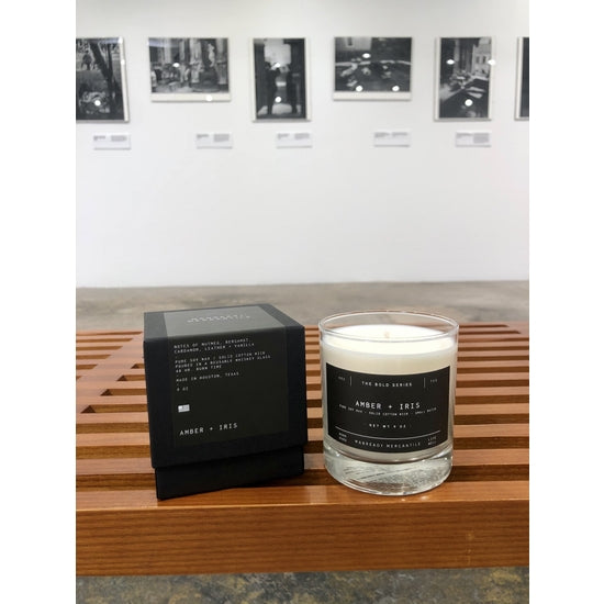Ace Rivington Manready Mercantile - Candle Amber + Iris placed on a wooden bench. The candle is in a clear glass container with a black label, and next to it is a black box with similar labeling. In the background, there is an art gallery wall displaying several black and white photographs.