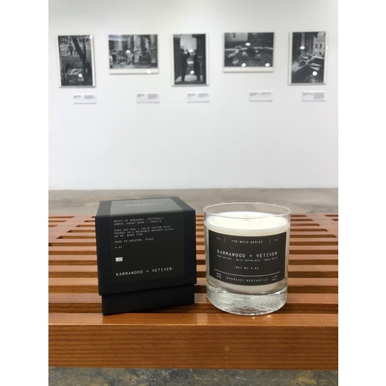 Ace Rivington Manready Mercantile - Candle Karmawood + Vetiver is displayed on a wooden bench. The candle is in a clear glass container with a gray label, and next to it is a black box with white text. In the background, there are several framed black-and-white photographs on a white wall, suggesting the setting is an art gallery or exhibition space. The candle and box are the main focus of the image.