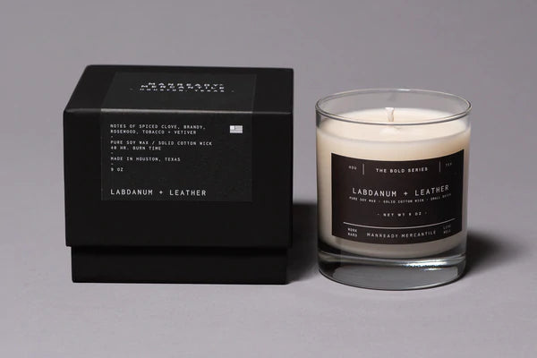 Ace Rivington Manready Mercantile - Candle Ladbanum + Leather. The candle, housed in a clear glass container with a black label, is situated next to its matching black packaging box. The label and packaging highlight the scent notes: spiced clove, brandy, mosswood, tobacco, and vetiver. The scene is set on a white background, emphasizing the candle's elegant and artisanal presentation.